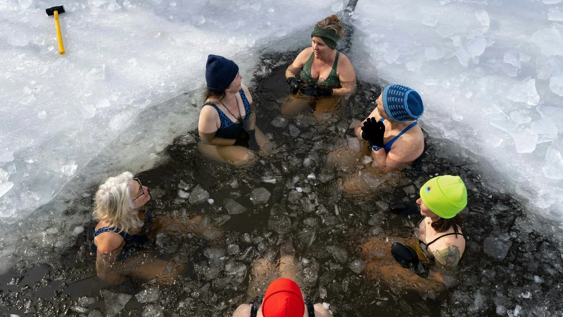 A group of women wearing winter hats and swimsuits sitting in a circular hole cut into a thick sheet of ice for cold-water plunging.