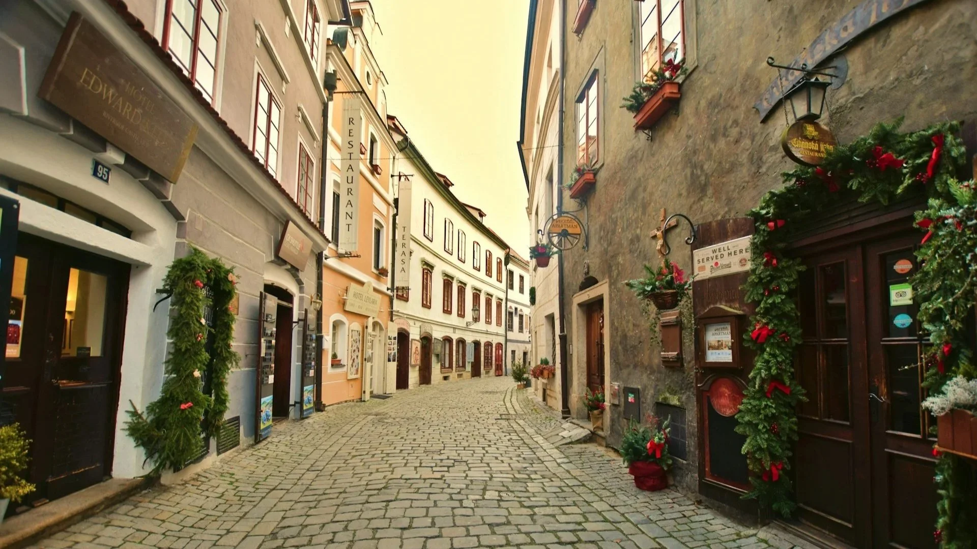 A narrow, winding cobblestone street lined with historic buildings decorated with evergreen garlands and festive wreaths.