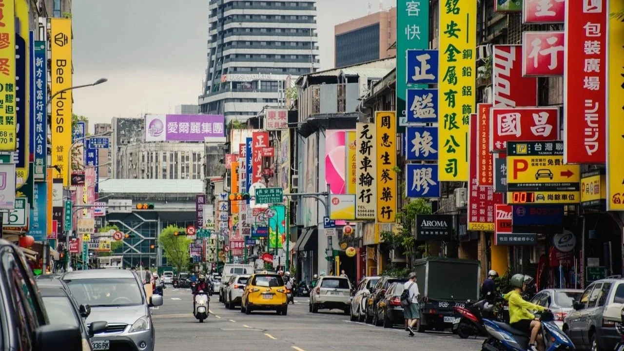A busy, multi-lane city street in Taiwan lined with vibrant, colorful vertical store signs in Chinese characters, featuring cars, scooters, and a tall modern skyscraper under an overcast sky in the background.