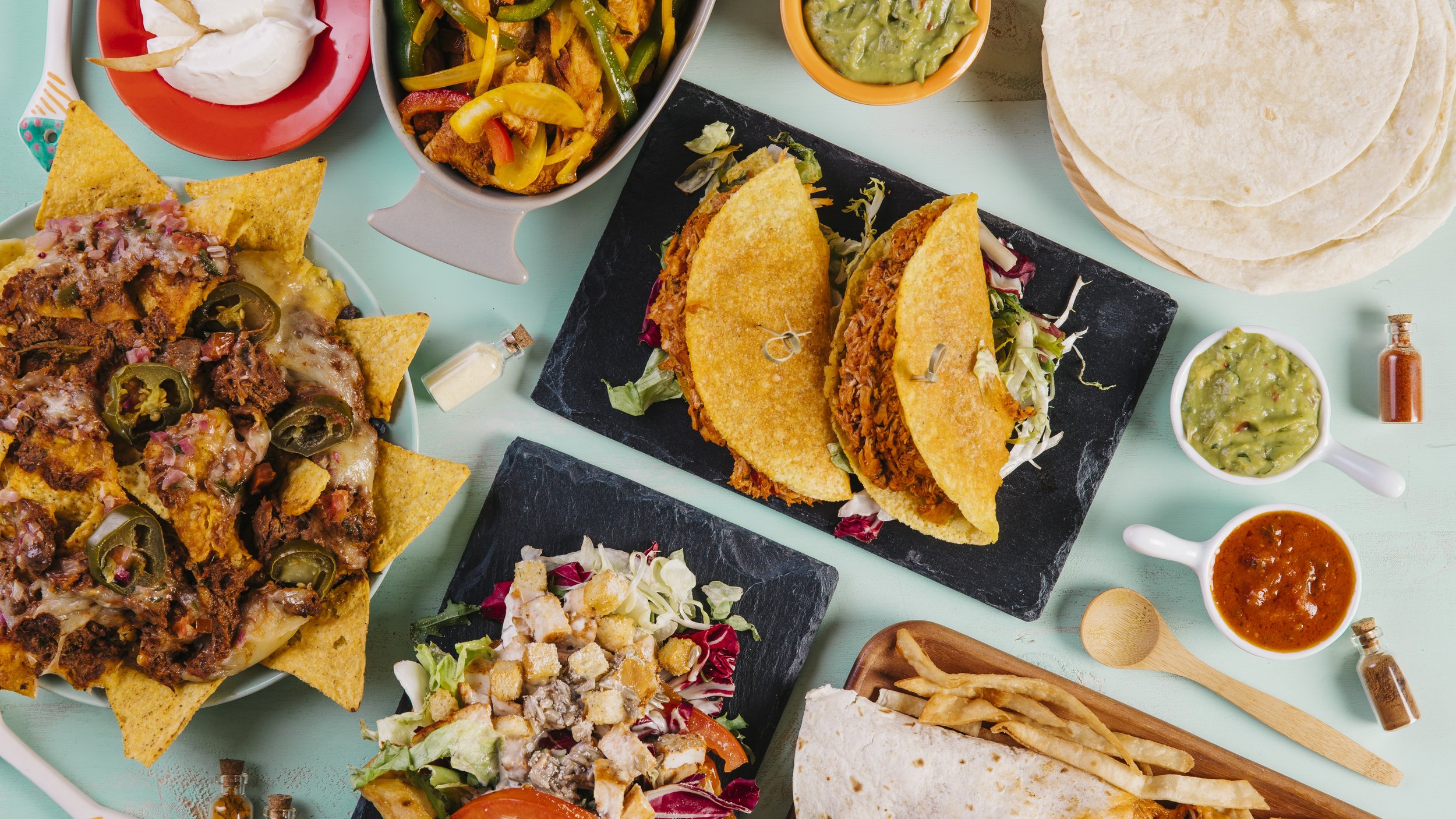 A vibrant spread of Mexican-style food, including nachos with jalapeños, hard-shell tacos, a salad, tortillas, and various bowls of salsa and guacamole.