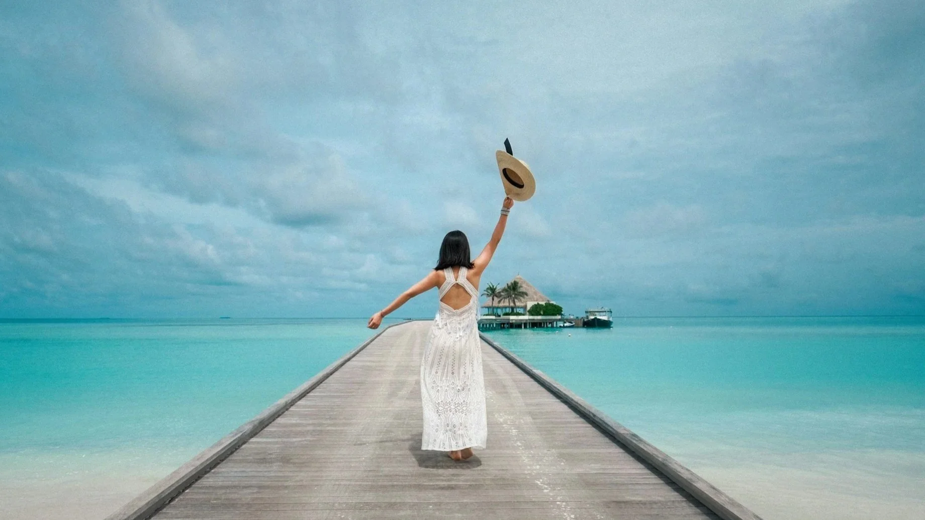 A woman in a white dress walks down a wooden pier toward a tropical hut, holding her hat up against a cloudy sky and calm turquoise sea.