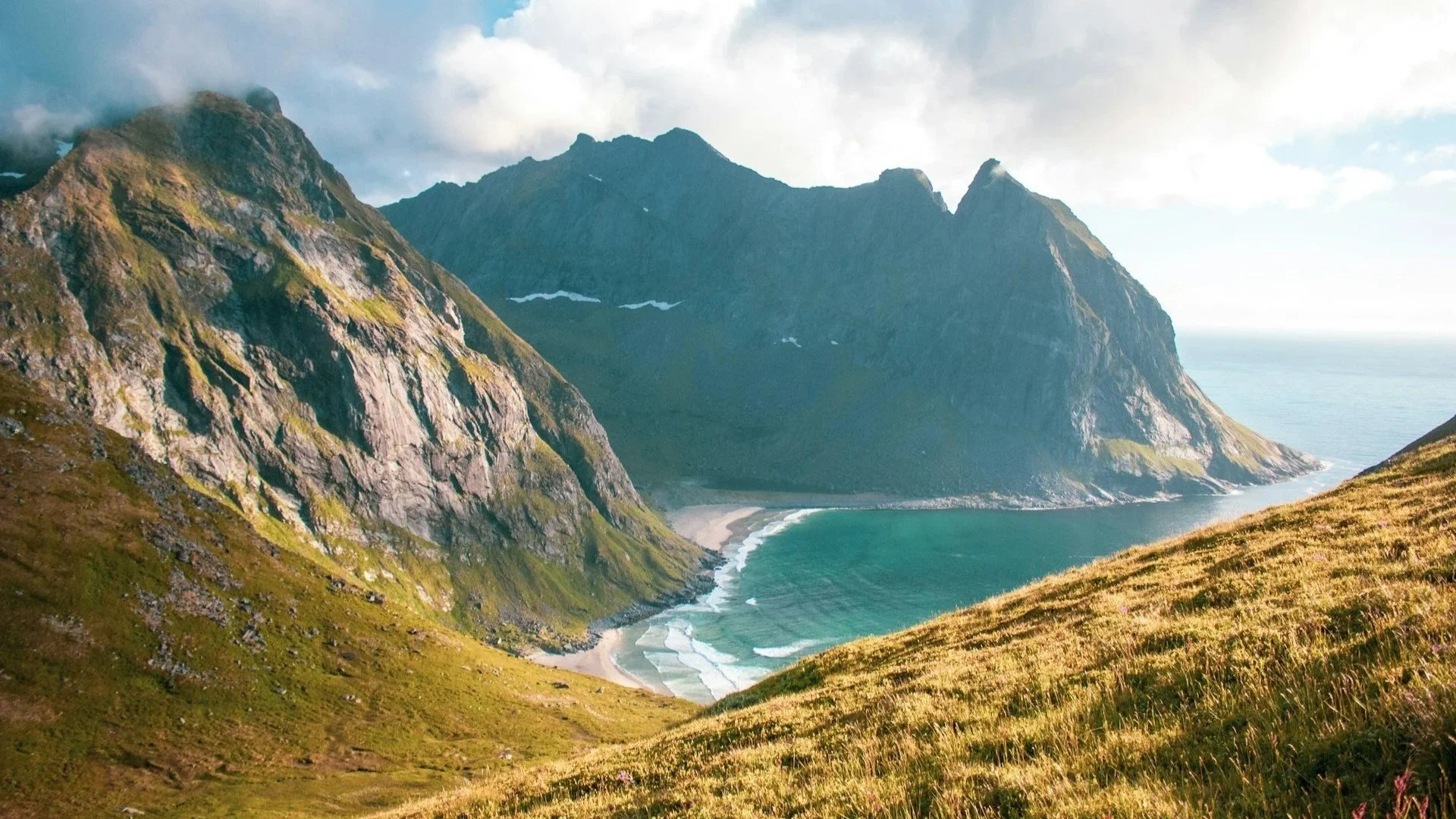 A wide-angle landscape of a turquoise bay nestled between steep, grassy mountains under a cloudy sky.