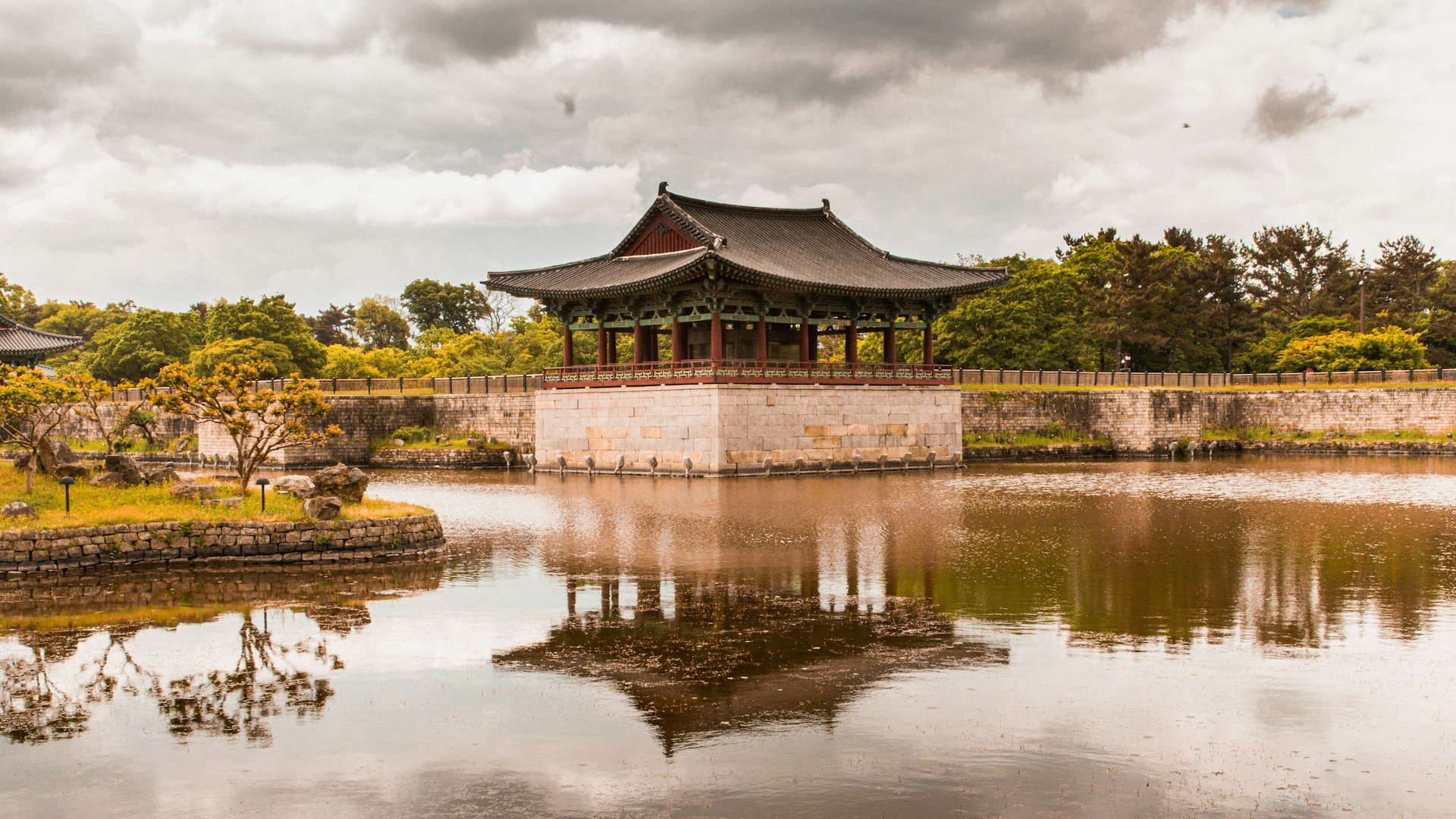 A classic Korean wooden pavilion with a tiled roof situated over a reflective pond under a cloudy sky.