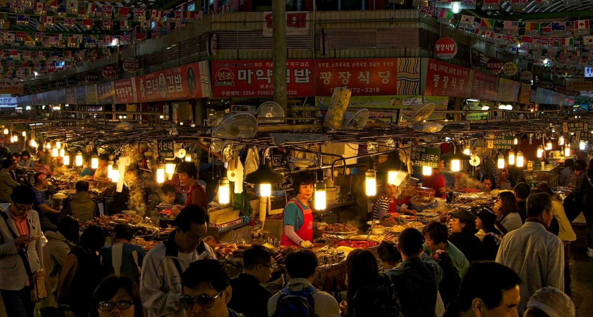 A busy, indoor night market with numerous food stalls under bright hanging lights, crowded with people and decorated with international flags.