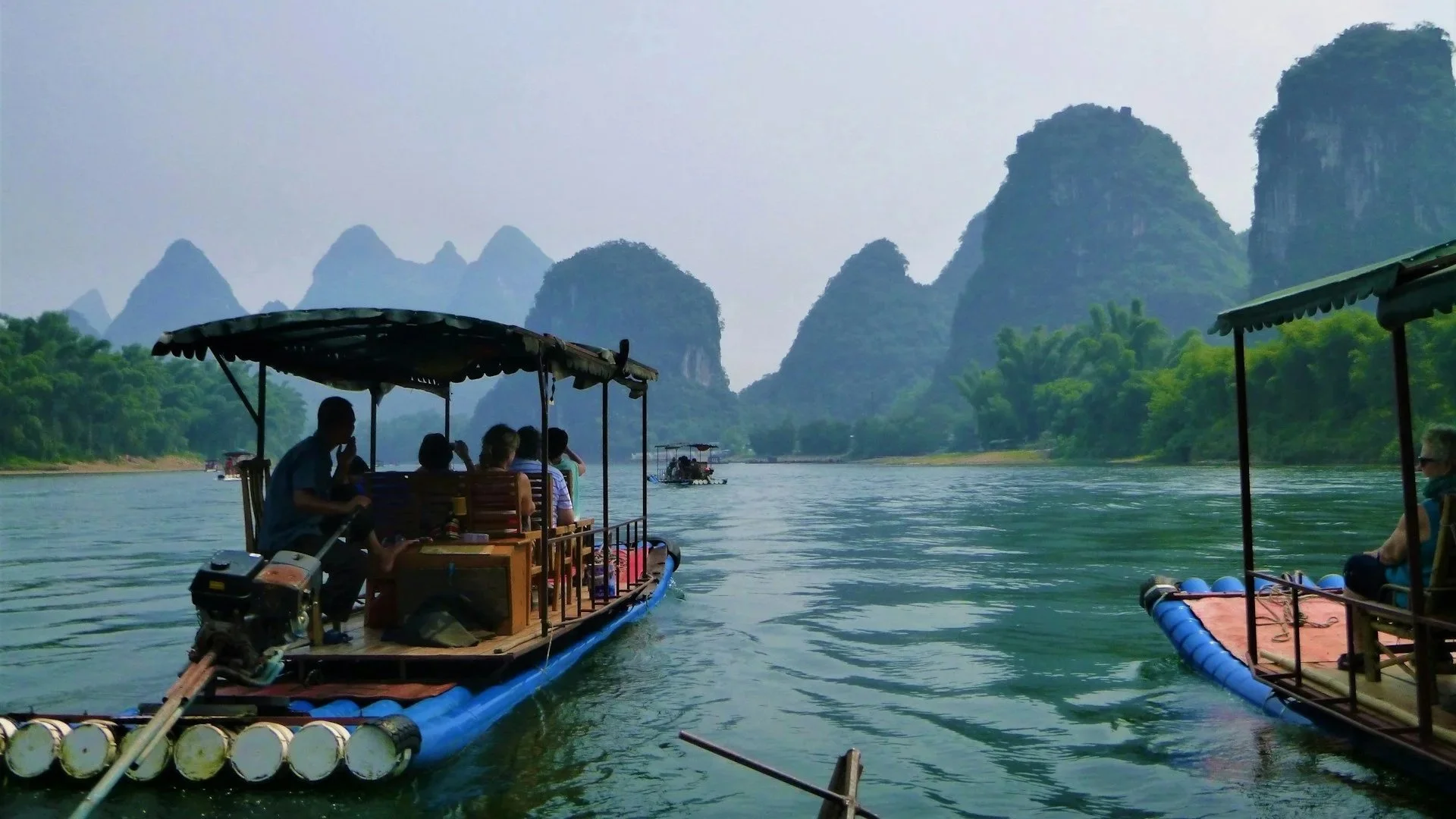 Passengers riding motorized bamboo rafts down a wide river surrounded by dramatic karst mountain peaks.