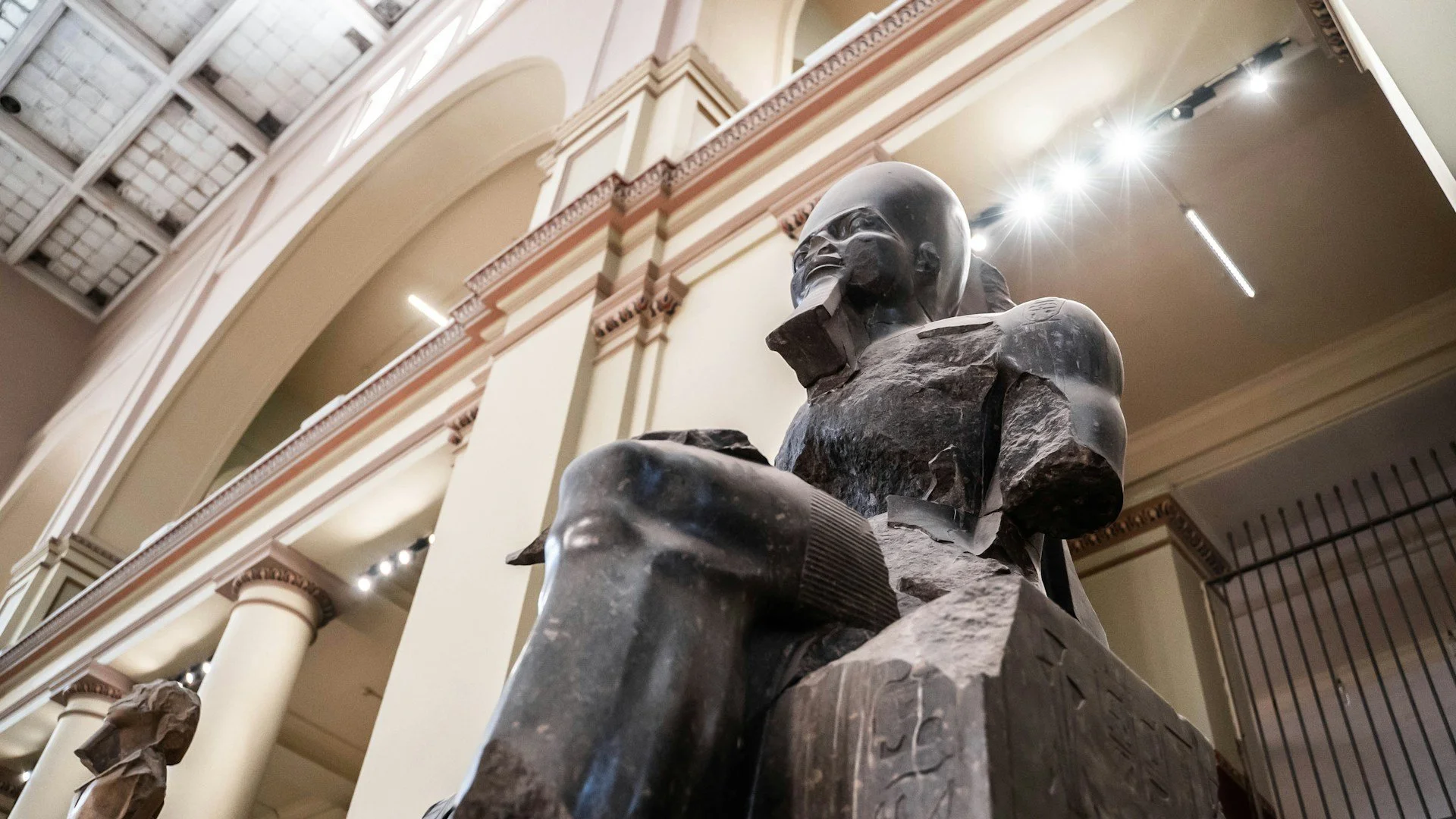 A low-angle shot of a colossal, seated black granite statue of an Egyptian pharaoh, displayed inside a grand museum with high arched ceilings and classical columns.