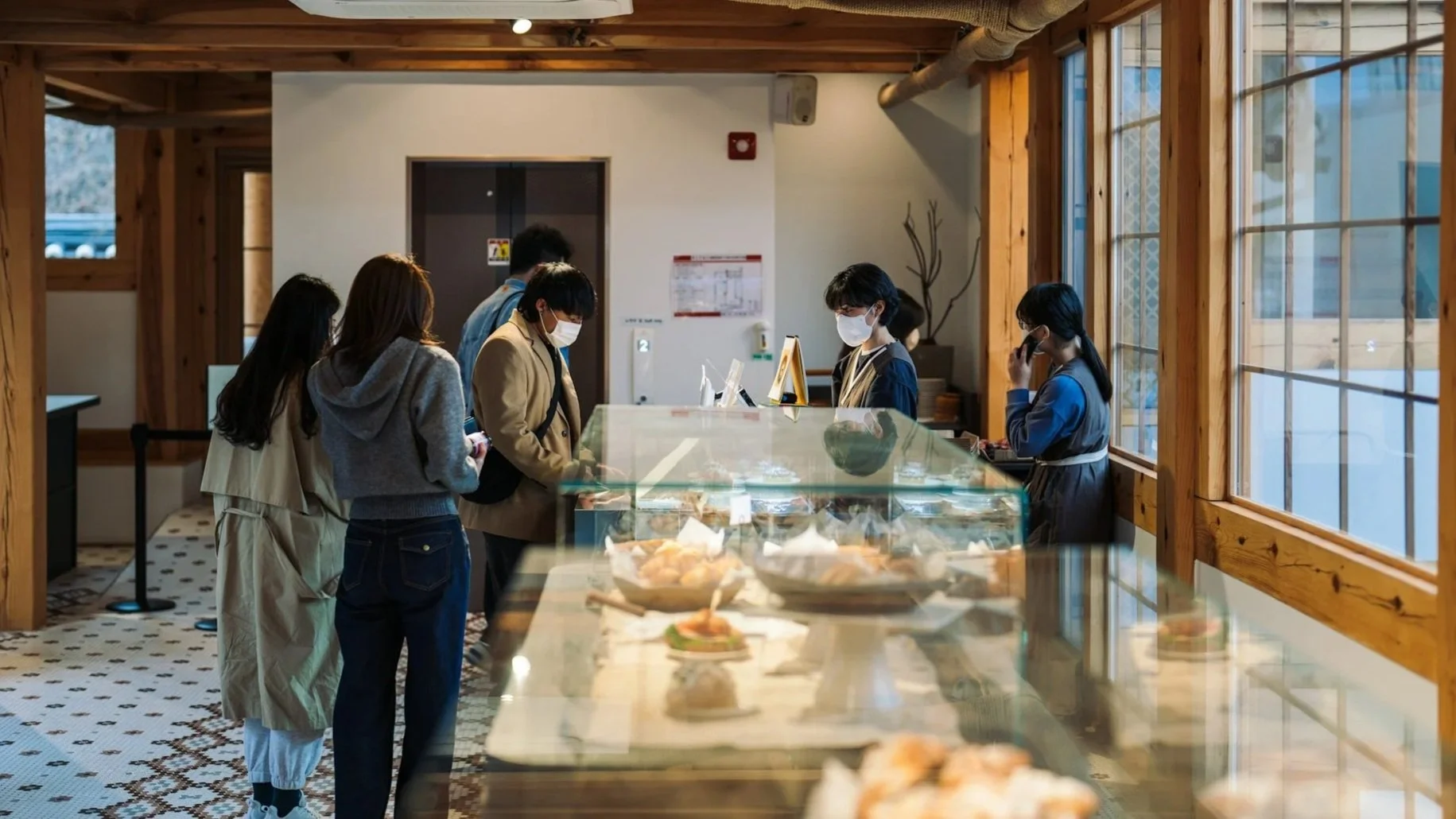 Customers wearing masks browse a glass display case of pastries inside a shop featuring traditional wooden post-and-beam architecture.