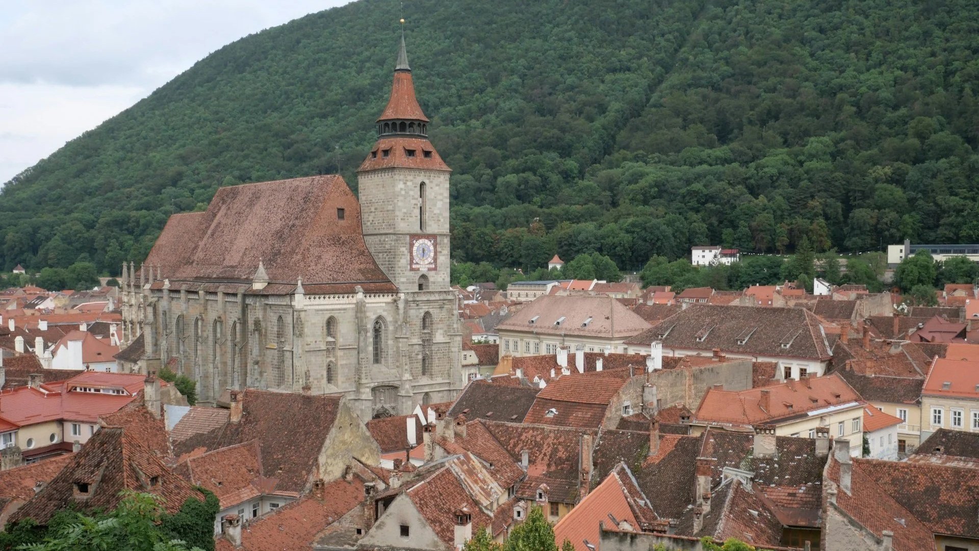The stone Black Church rising above a dense cluster of historic terracotta tiled rooftops in Brașov, with a forested hill in the background.