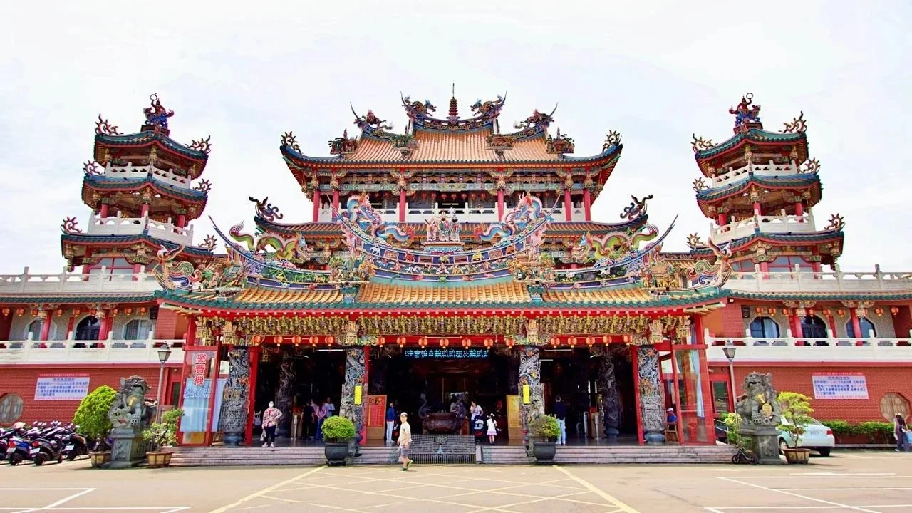 A wide front view of a large traditional Taiwanese temple complex under a bright sky, featuring multi-tiered swooping roofs heavily decorated with colorful, intricate dragon sculptures and ornate carvings above the main entrance.