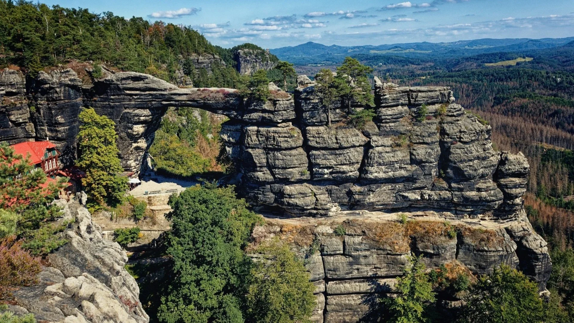 A large natural sandstone arch bridge surrounded by green forest and a red-roofed building in the Bohemian Switzerland National Park.