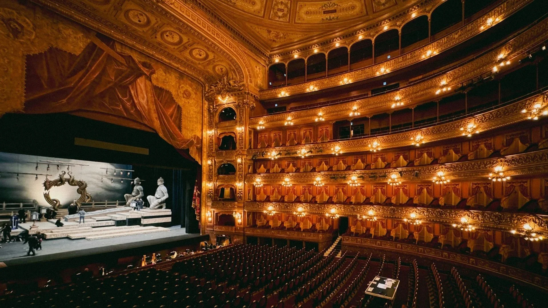 A wide view of a grand, ornate opera house interior with multiple tiers of golden, illuminated balconies overlooking a stage set with large statues.