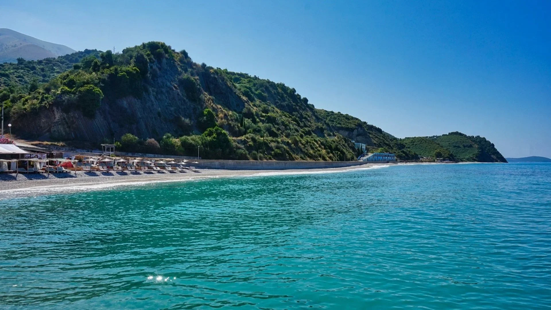 Clear turquoise sea along a pebbled beach lined with sun loungers against green hills.