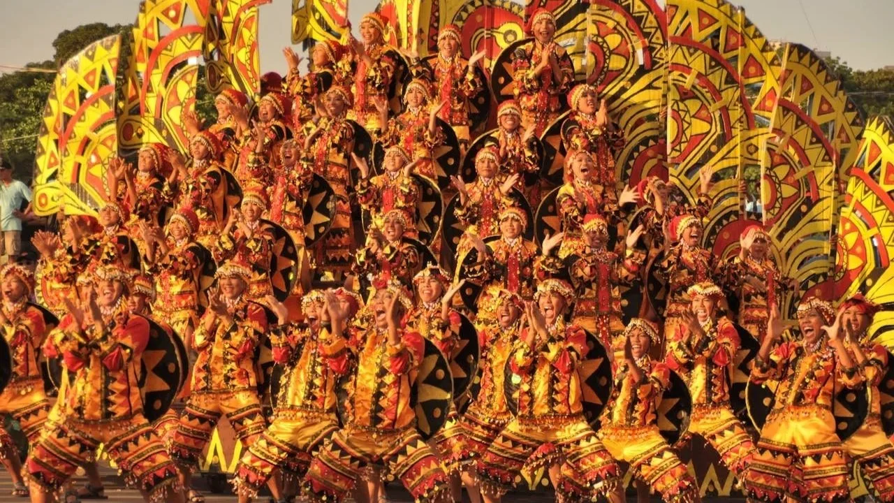 A large group of festival dancers performing on a street, wearing highly detailed, matching yellow, orange, and black patterned costumes and headpieces.
