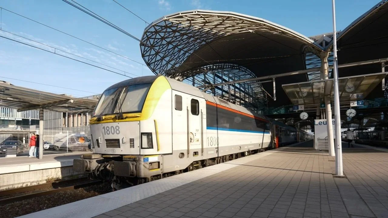 A passenger train parked at a modern, covered Belgian railway station platform on a sunny day.
