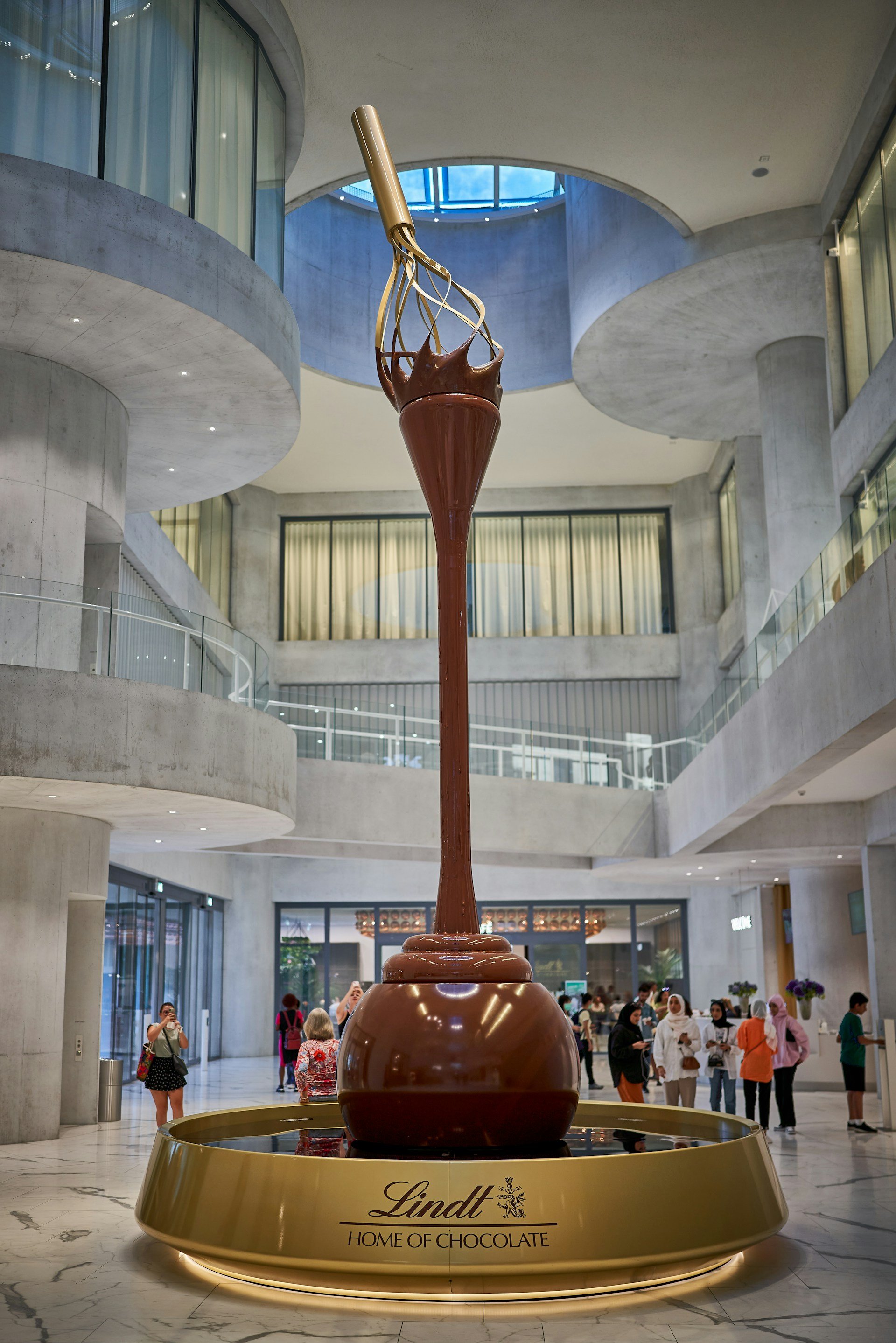 The interior of the Lindt Home of Chocolate in Zurich, featuring a massive, multi-story chocolate fountain shaped like a giant golden whisk dripping with liquid chocolate.