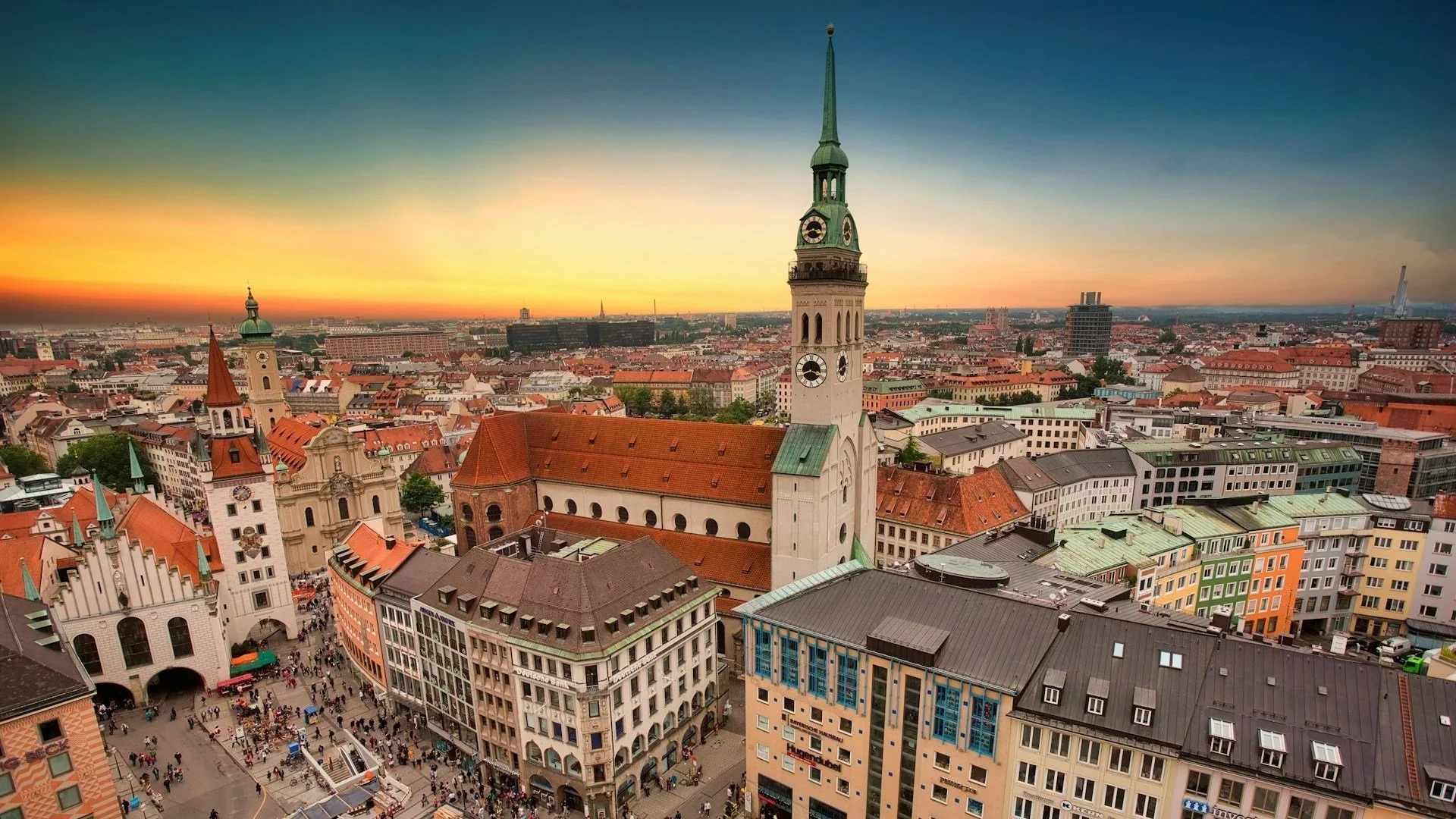 High-angle shot of Marienplatz in Munich, Germany, looking down at bustling pedestrian crowds and featuring the prominent, tall green spire of St. Peter's Church against a vibrant sunset.