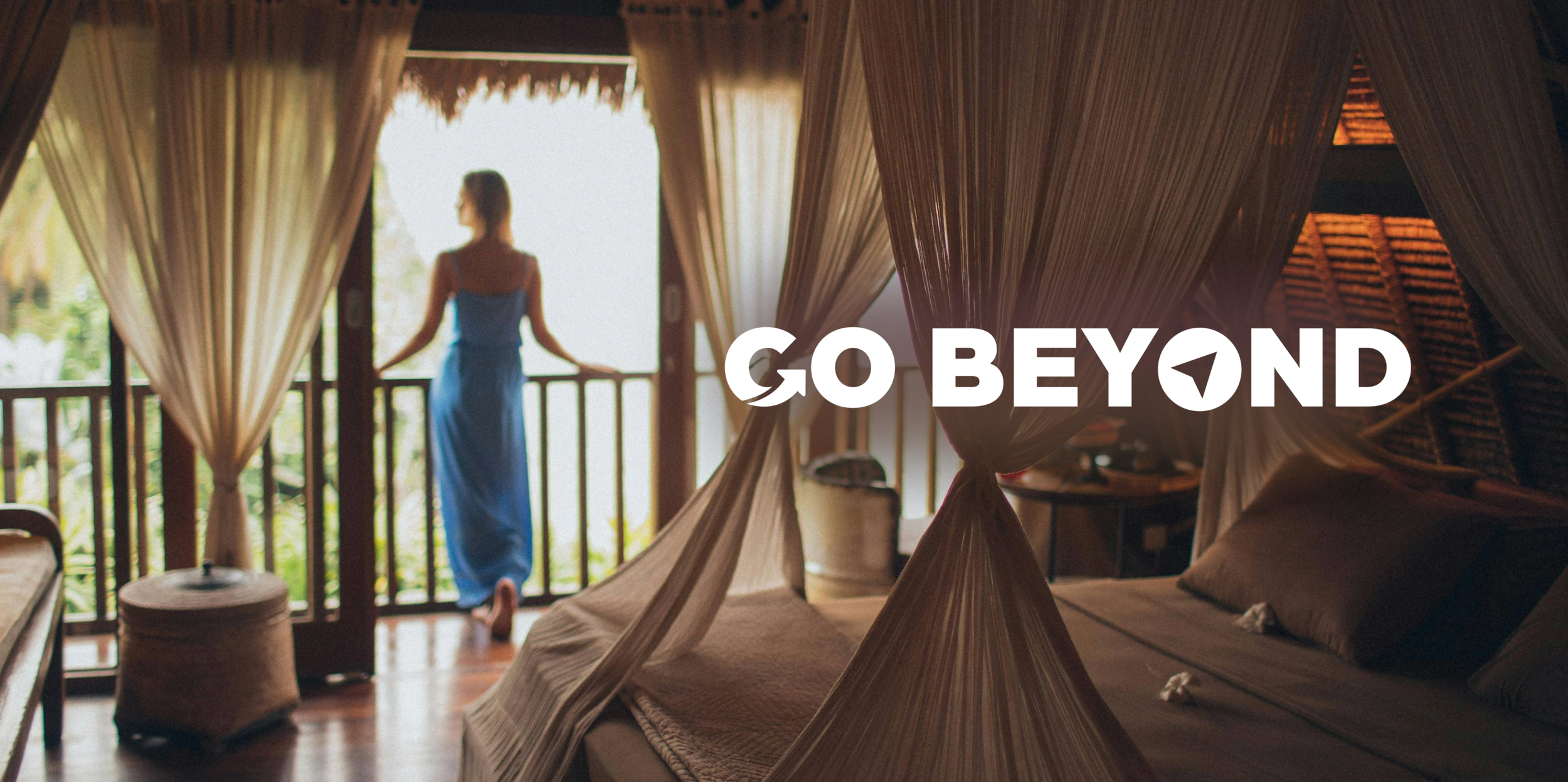 A woman in a blue dress stands on a balcony, looking out at a tropical view. She is in a rustic room with a large bed with a canopy.