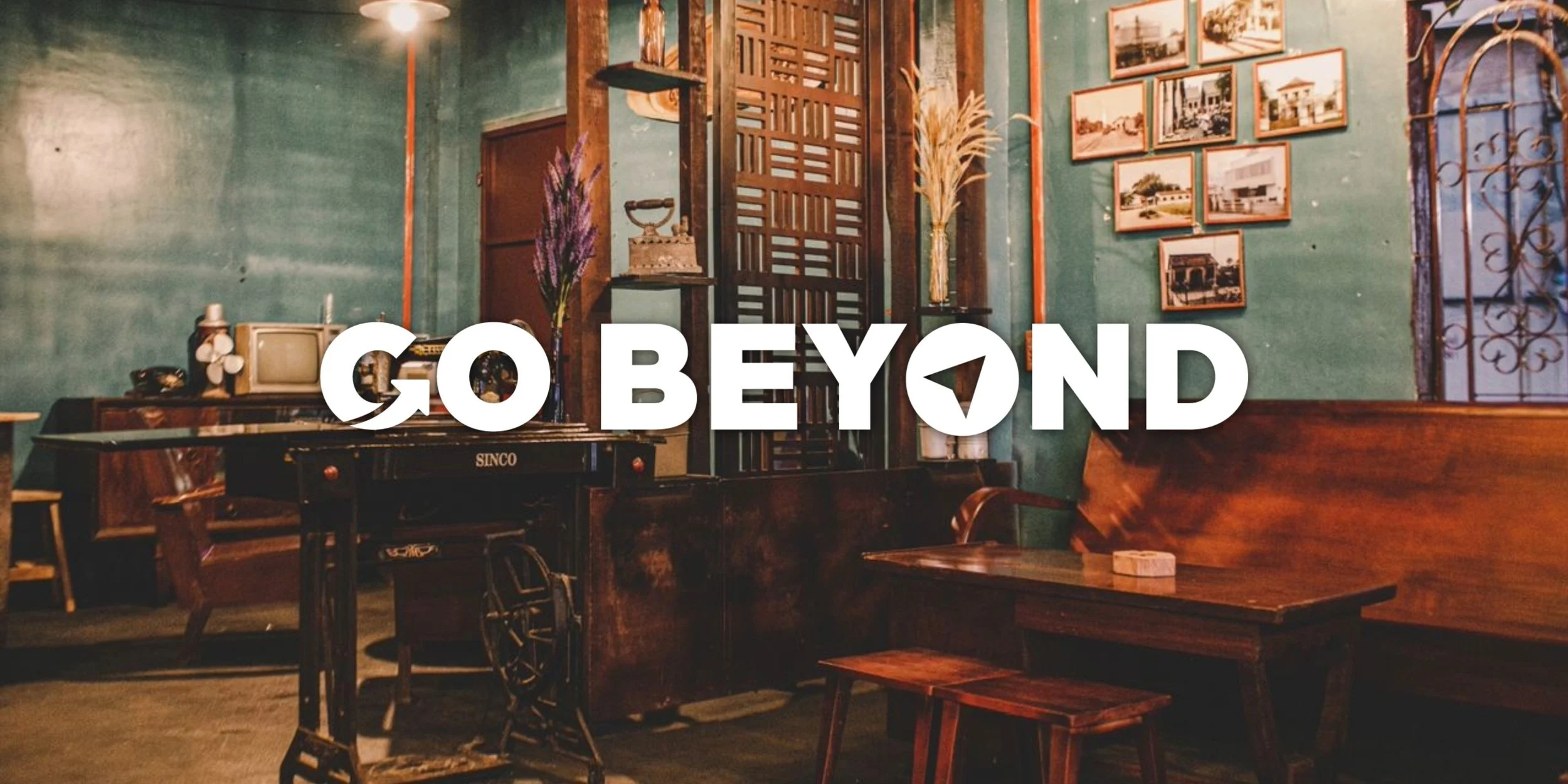 Vintage-style interior featuring retro wooden furniture, framed sepia photographs on teal walls, and an old sewing machine, overlaid with white text reading "GO BEYOND."
