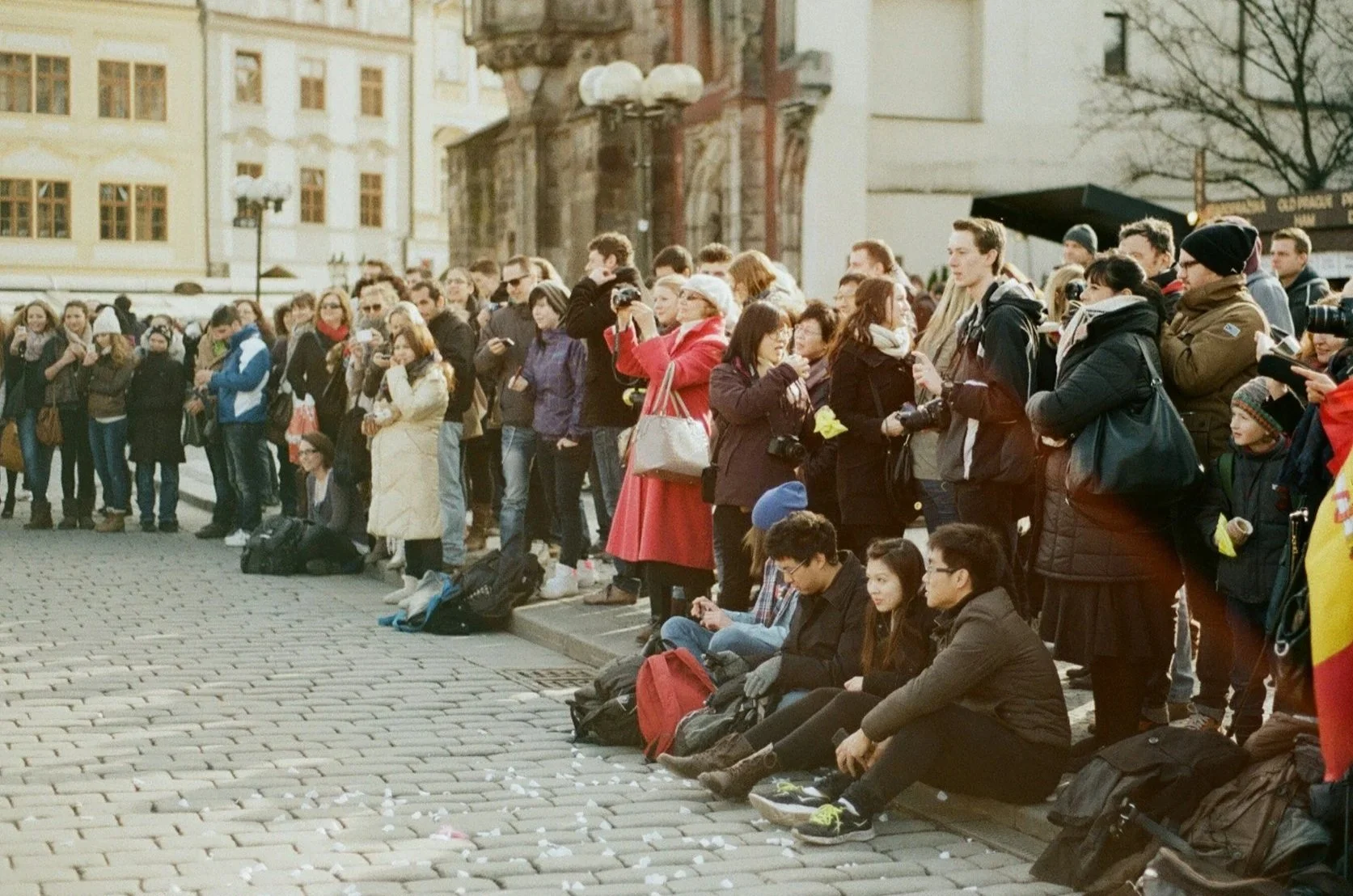 A large, diverse crowd of tourists standing and sitting on a cobblestone square in front of historic European buildings.