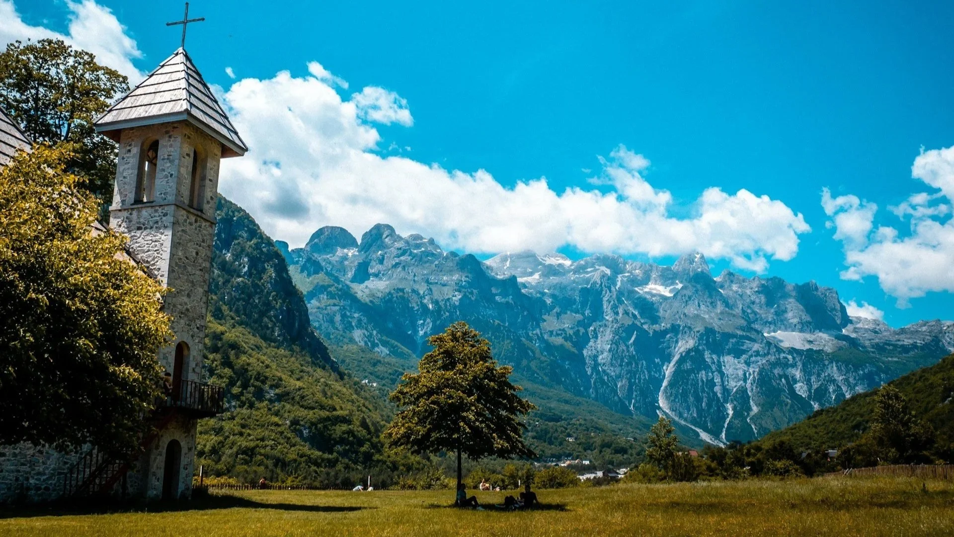 Historic stone church tower on a grassy field with dramatic, jagged mountains under a blue sky.