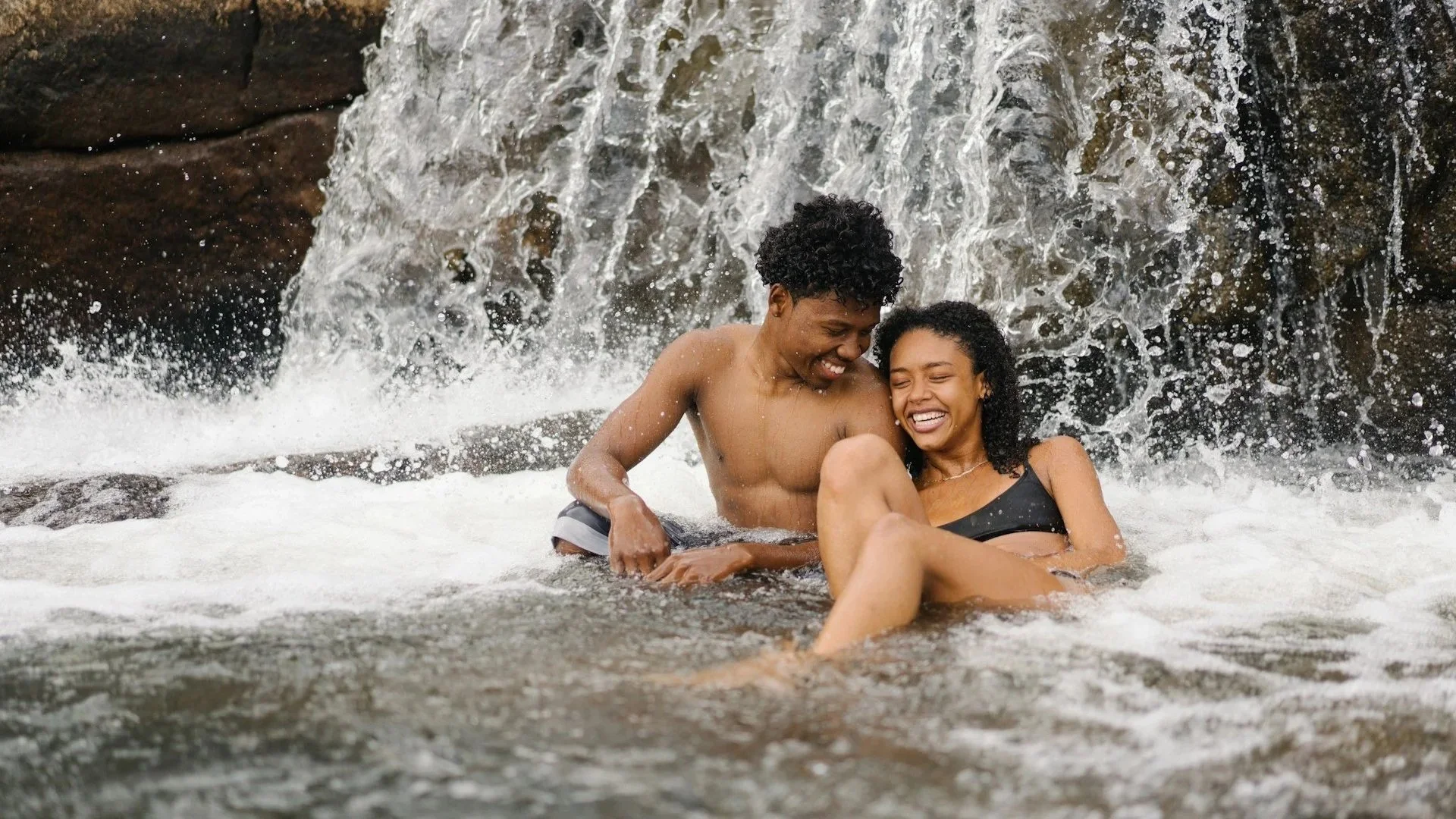 A young Black couple laughs and smiles while sitting together in the rushing water at the base of a rocky waterfall.