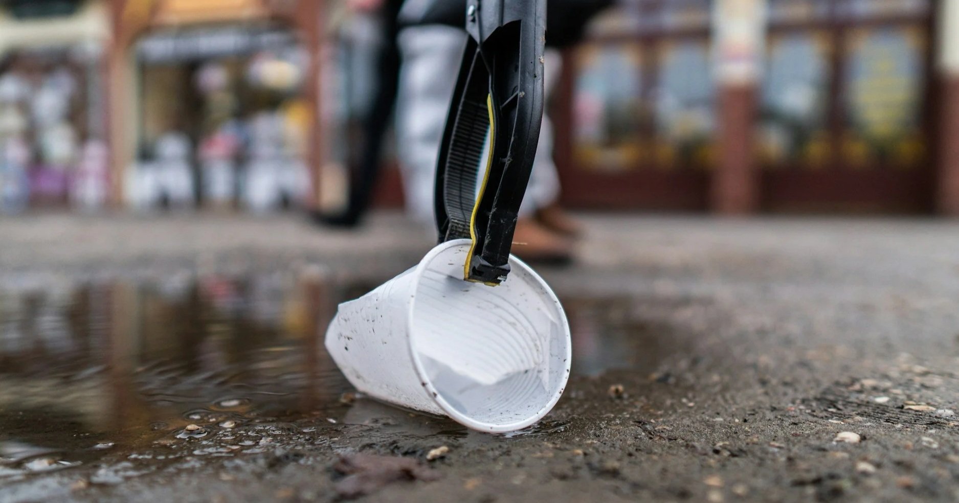 A close-up, low-angle shot of a litter-picker tool grasping a white plastic cup from a puddle on a city street.