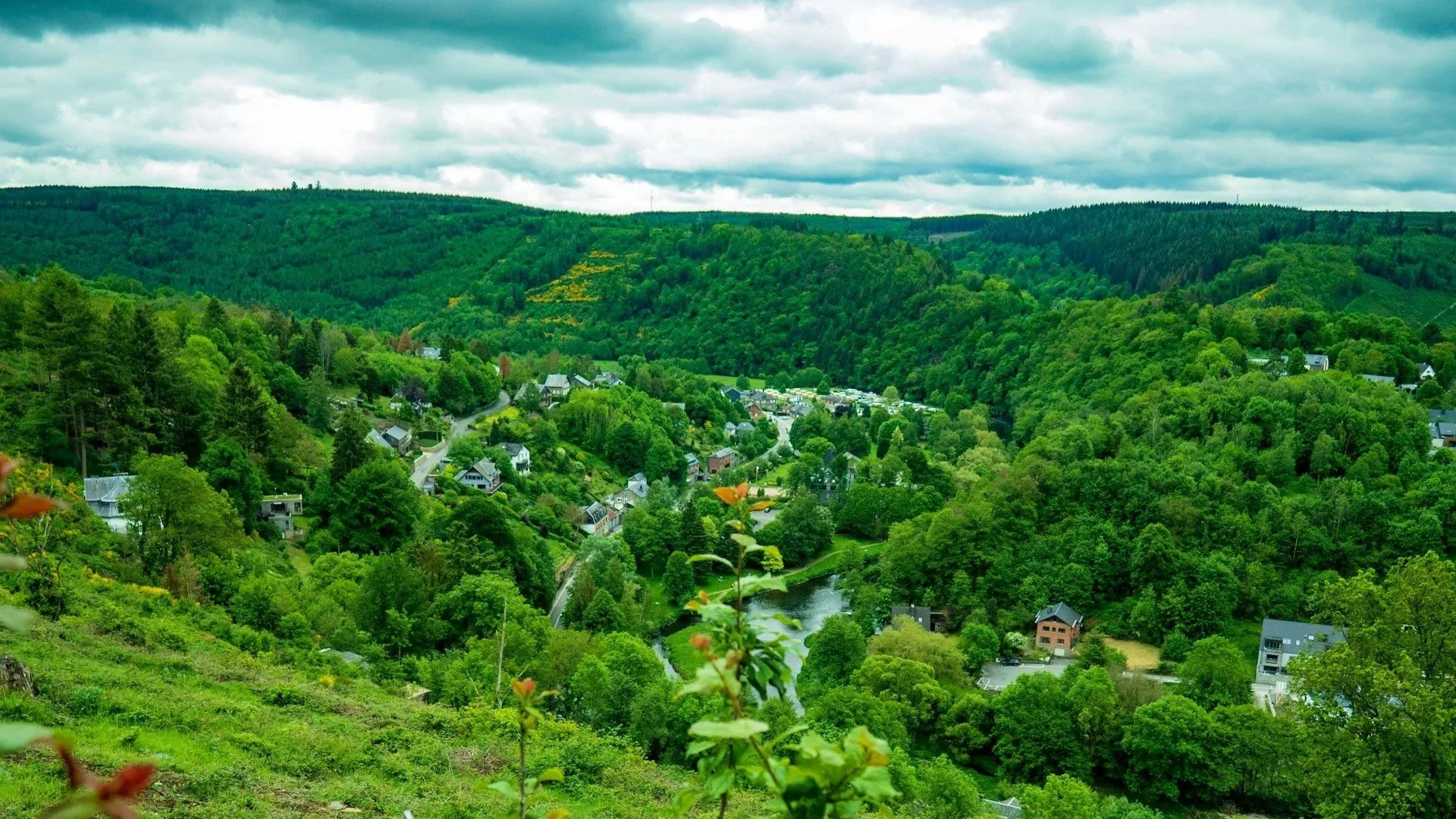 High-angle view of a lush green valley in the Belgian Ardennes with small village houses nestled along a winding river.