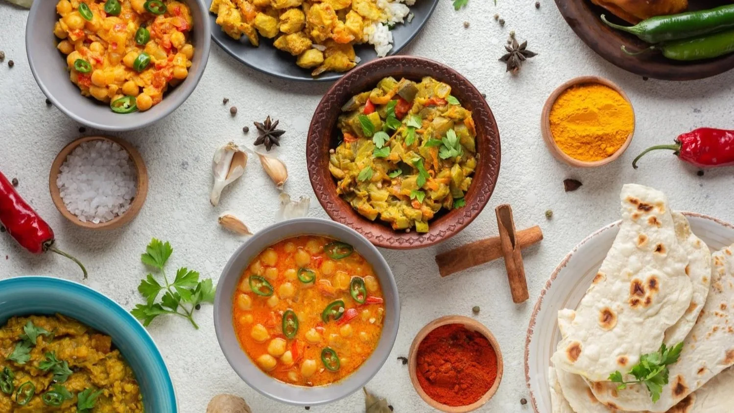 Top-down view of an Indian food spread featuring various bowls of curry, chickpeas, naan bread, and spices.