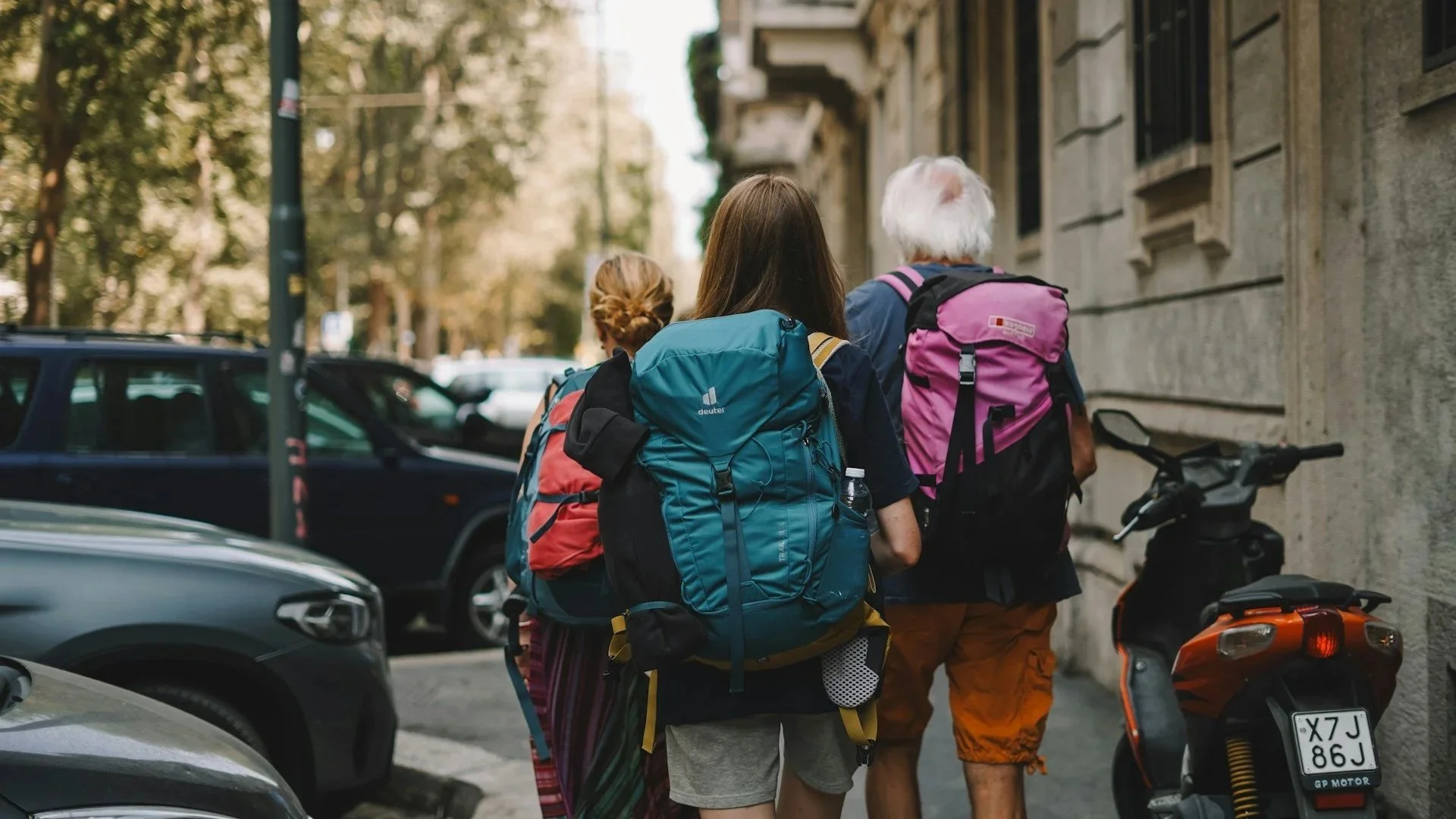 A rear view of three people wearing large hiking backpacks as they walk down a narrow city street lined with parked cars and a moped.