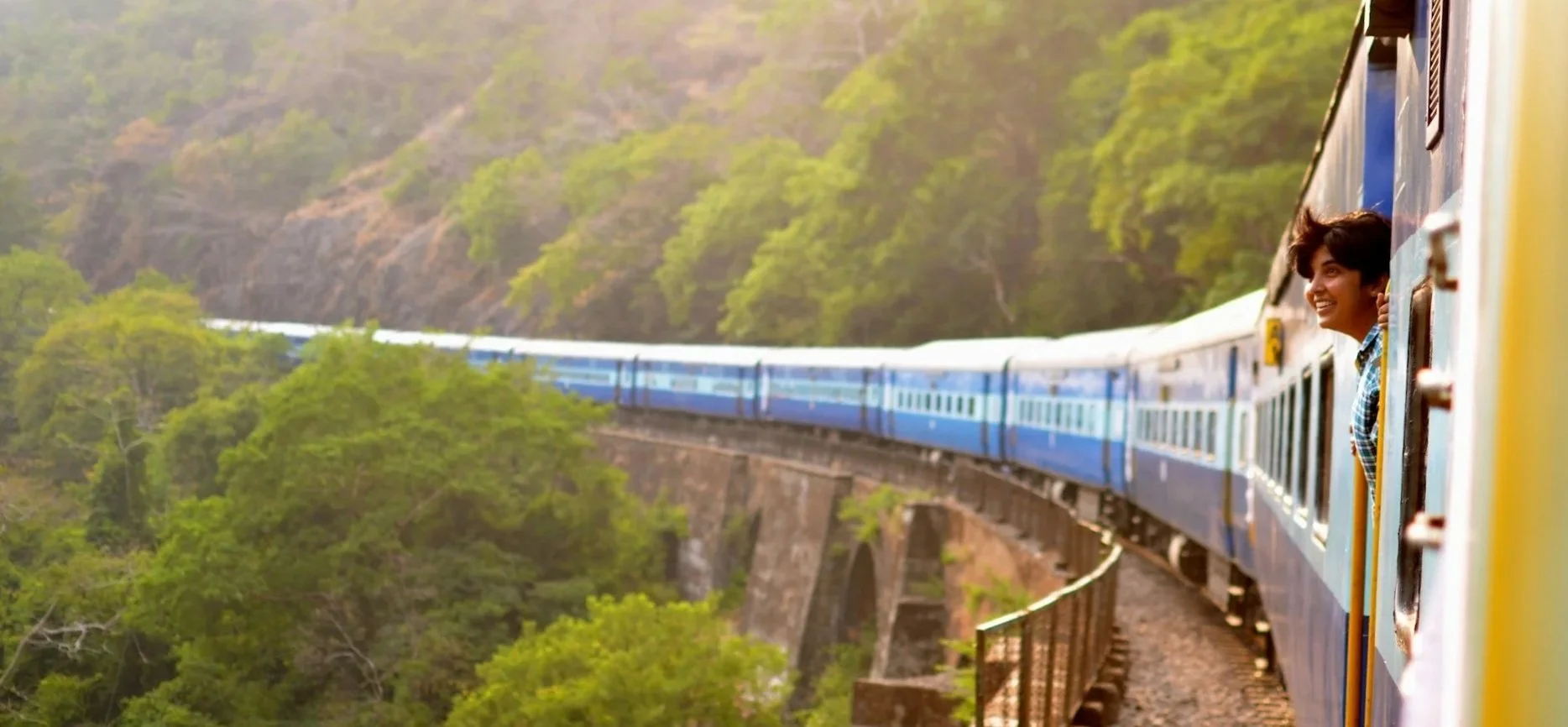 A smiling traveler leaning out of a blue passenger train window as it curves through a sunlit, lush green valley.