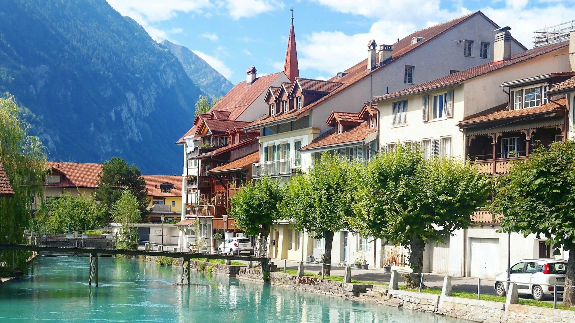 Traditional Swiss buildings with red-tiled roofs lining a bright turquoise canal, framed by red flowers in the foreground and steep mountains behind.