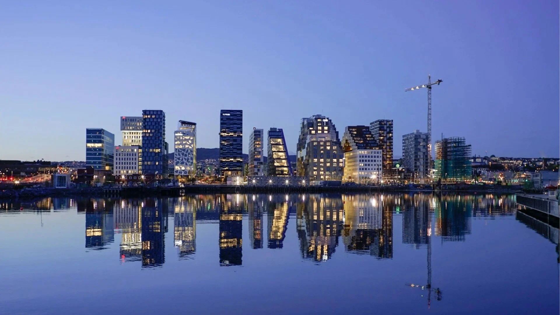 Gemini said A night skyline of the Barcode Project in Oslo, Norway, featuring modern high-rise buildings illuminated and reflecting on the calm surface of a dark blue harbor.