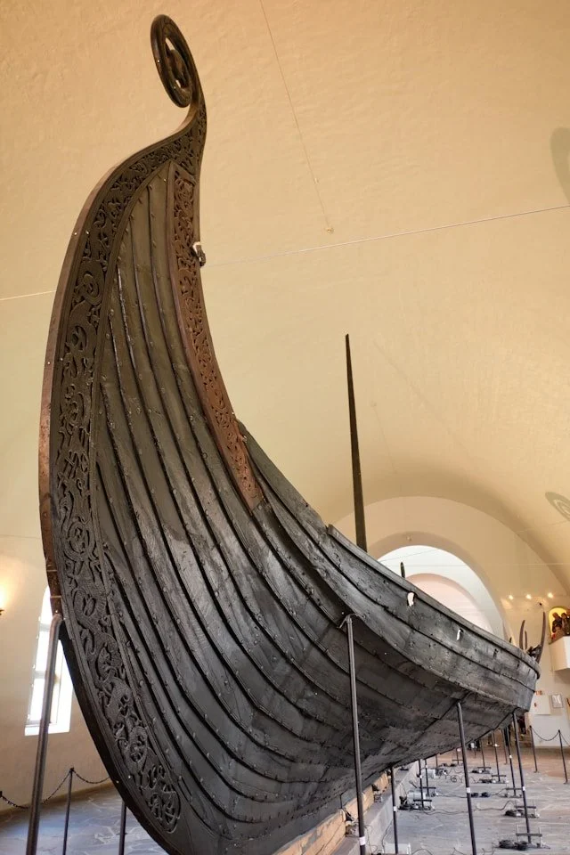 A close-up, low-angle shot of the Oseberg Viking ship’s prow, featuring dark, weathered wood and elaborate serpent-like carvings, displayed inside a museum with white vaulted ceilings.