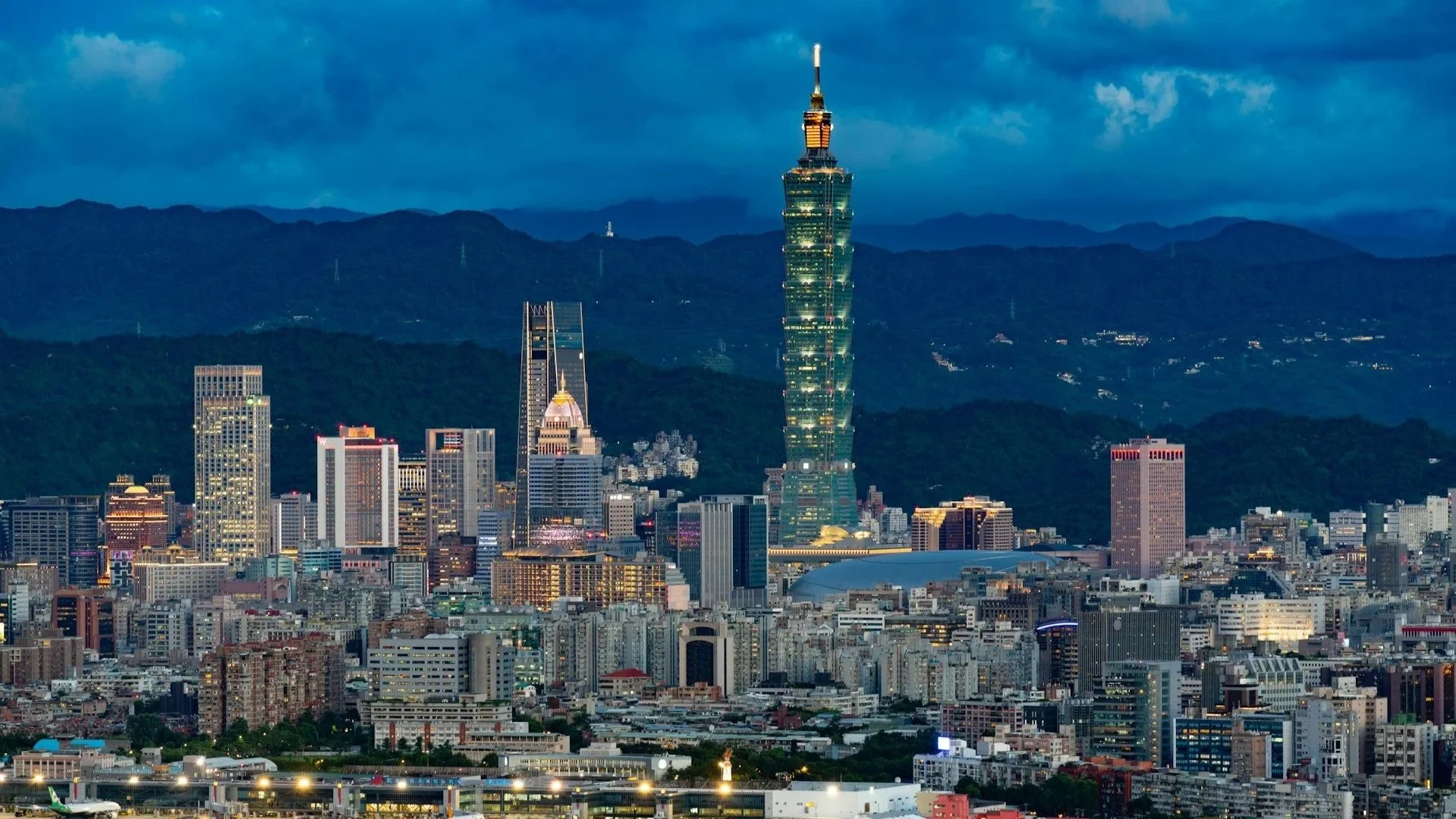A wide city skyline at twilight featuring the brightly lit Taipei 101 skyscraper against a backdrop of dark mountains and a deep blue evening sky.
