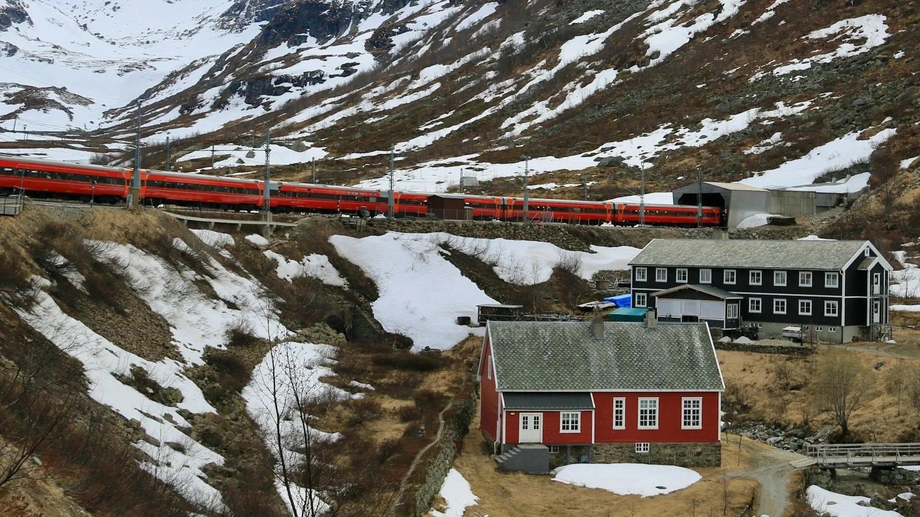 A long red train travels across a snowy mountain landscape in Norway, passing behind a traditional red wooden house and a larger black building nestled in a valley.