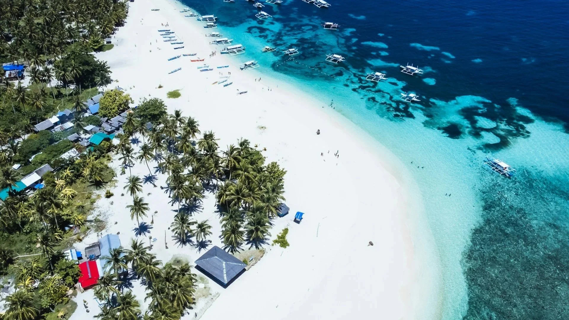 Aerial view of a wide white-sand beach with palm trees, clear turquoise water, and traditional outrigger boats anchored near the shore.