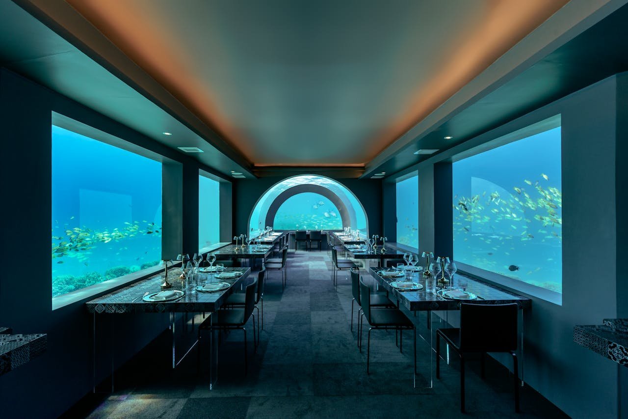 An elegant, dimly lit underwater restaurant featuring large viewing windows that reveal schools of yellow fish swimming in clear blue water.