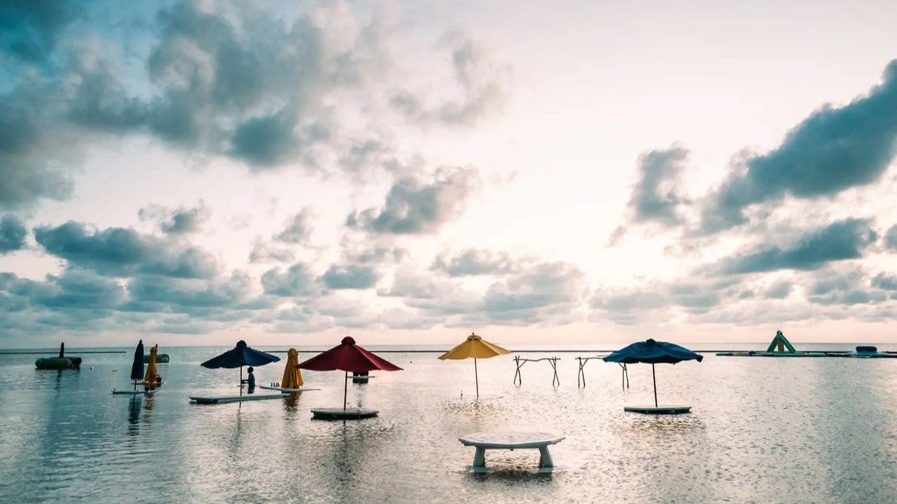 Colorful beach umbrellas and tables stand directly in calm, shallow ocean water under a cloudy, sunset sky.