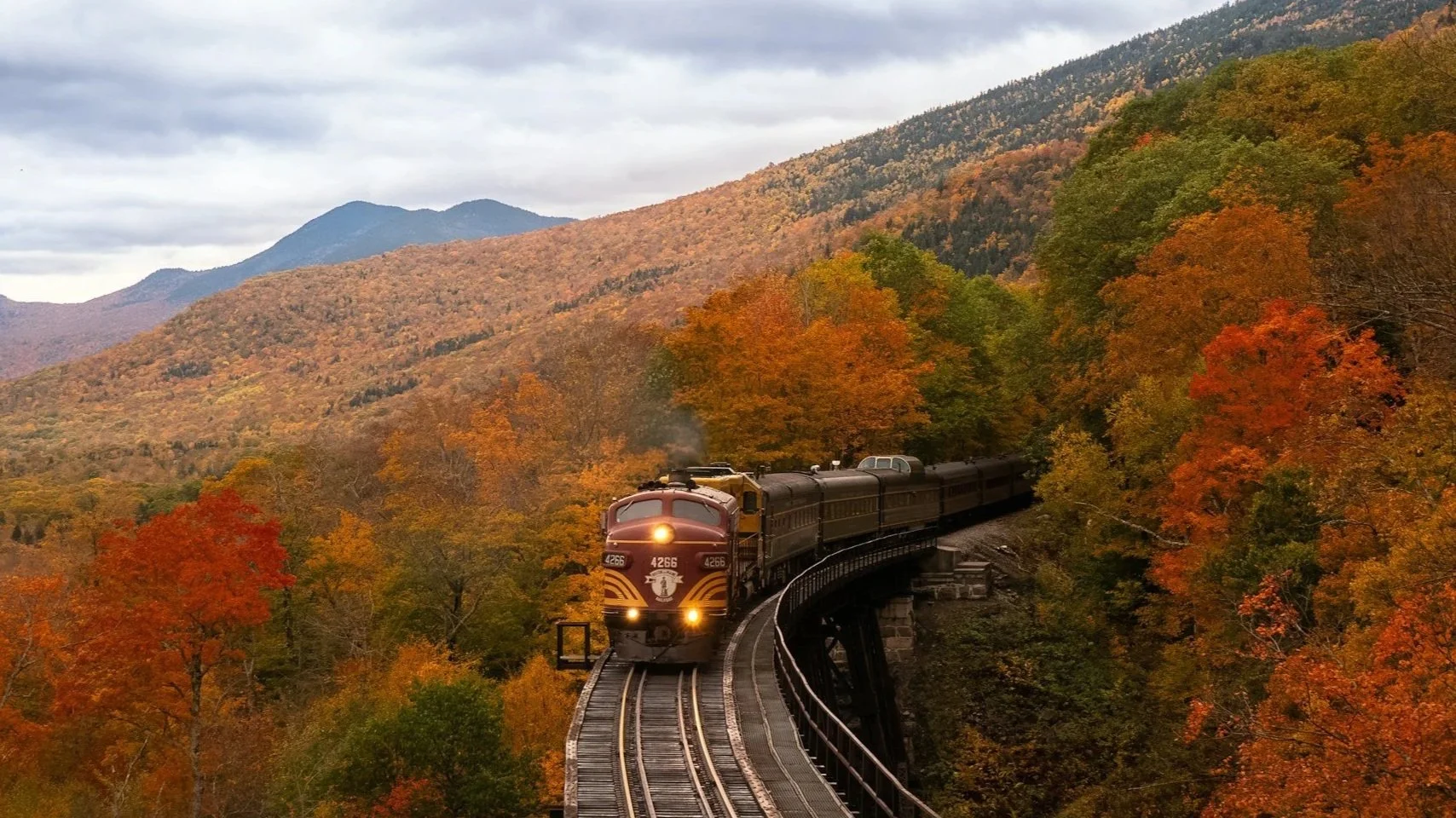 A classic passenger train traveling across a high wooden trestle bridge, surrounded by vibrant autumn trees in shades of orange, red, and yellow with mountains in the background.