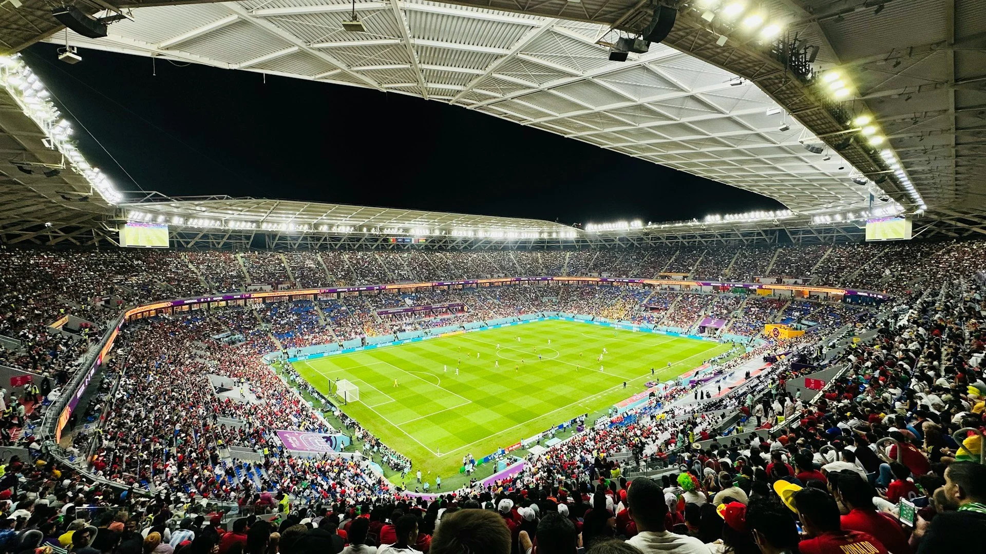 A wide-angle, high-angle view of a packed football stadium at night under bright floodlights during a match.