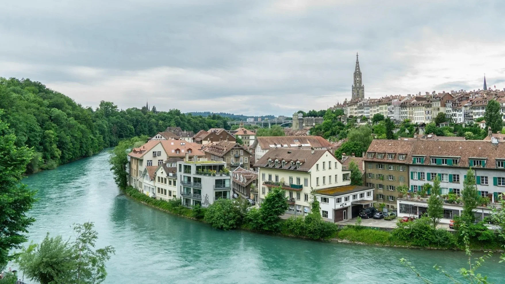 A view of Bern's old town featuring historic buildings with red-tiled roofs along a turquoise river, with a tall church spire rising in the distance.