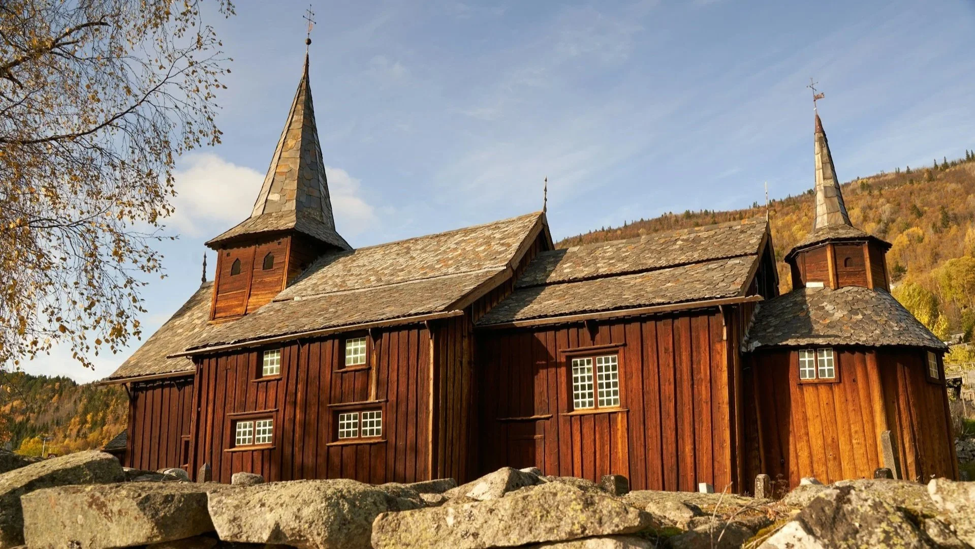 A side-profile view of the Nore Stave Church in Norway, featuring dark timber walls and two pointed stone-shingled spires, seen from behind a rustic stone wall under a clear blue sky.