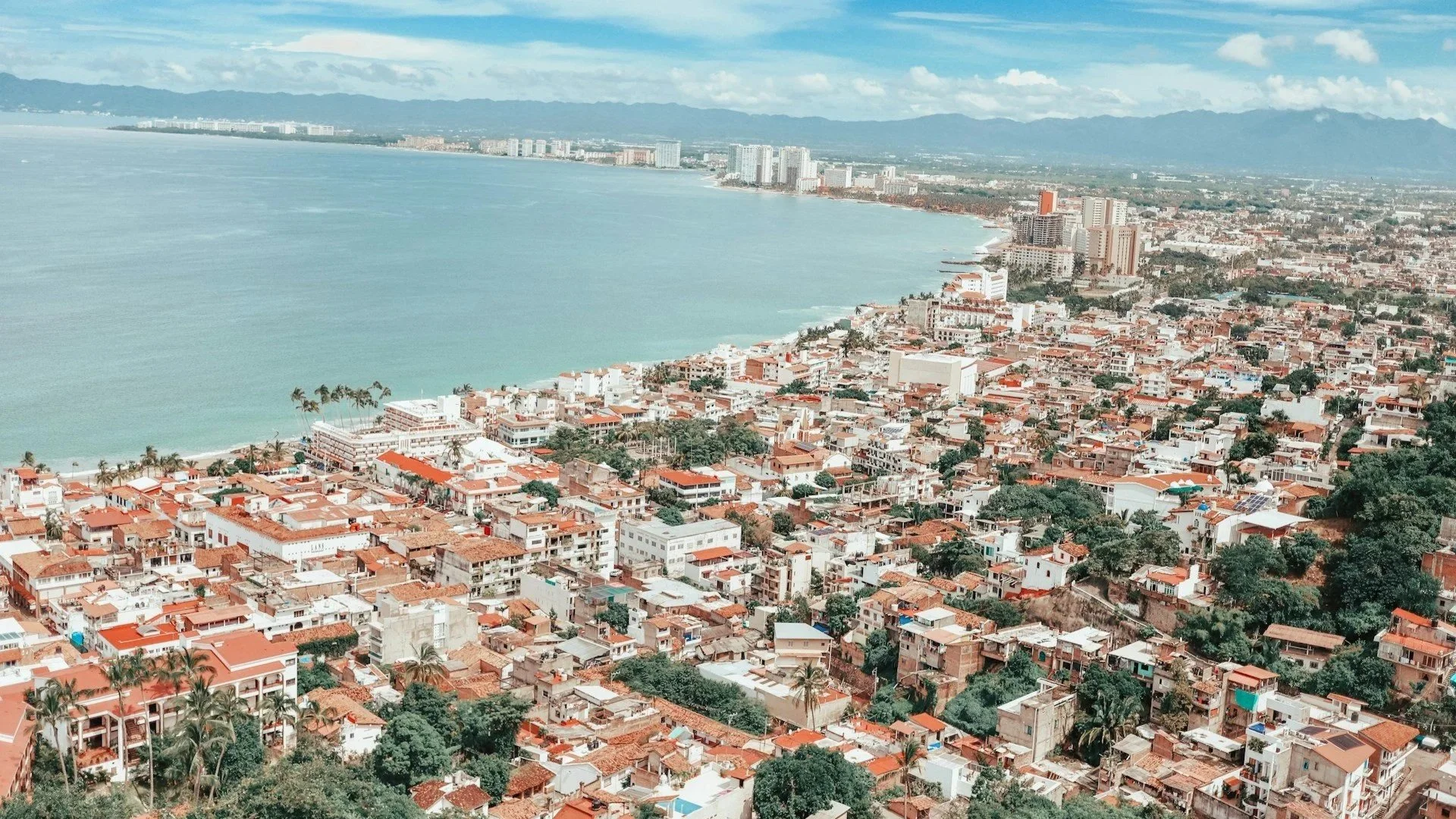 An elevated view of a coastal city with white and red-roofed buildings densely packed along a curved bay with blue water and distant mountains.