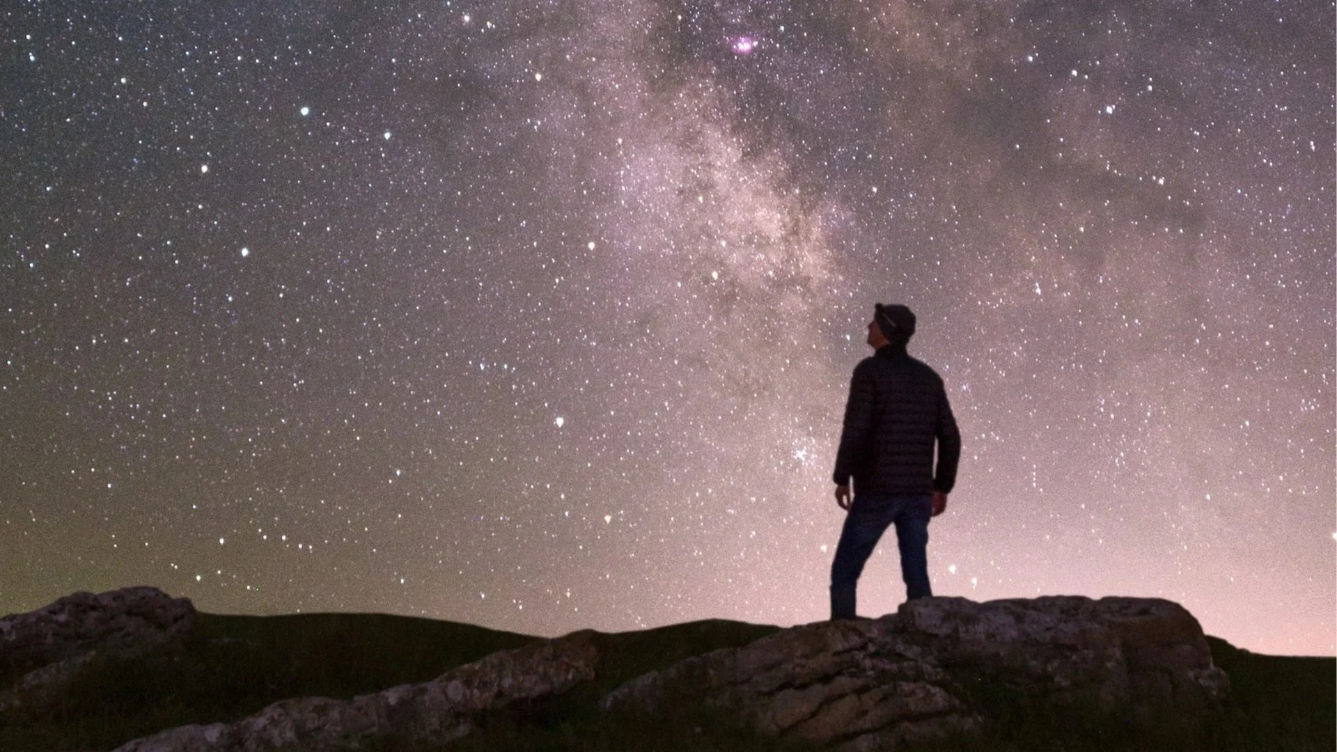 A silhouette of a person standing on a rocky hill, looking up at the vast, glowing Milky Way galaxy in a dark night sky.