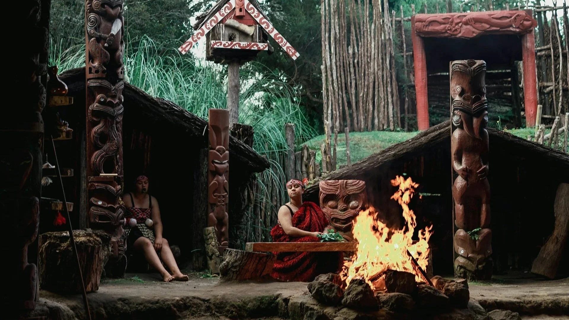 A traditional Māori village scene featuring carved wooden figures (Pou), people in traditional dress, and a roaring central fire.