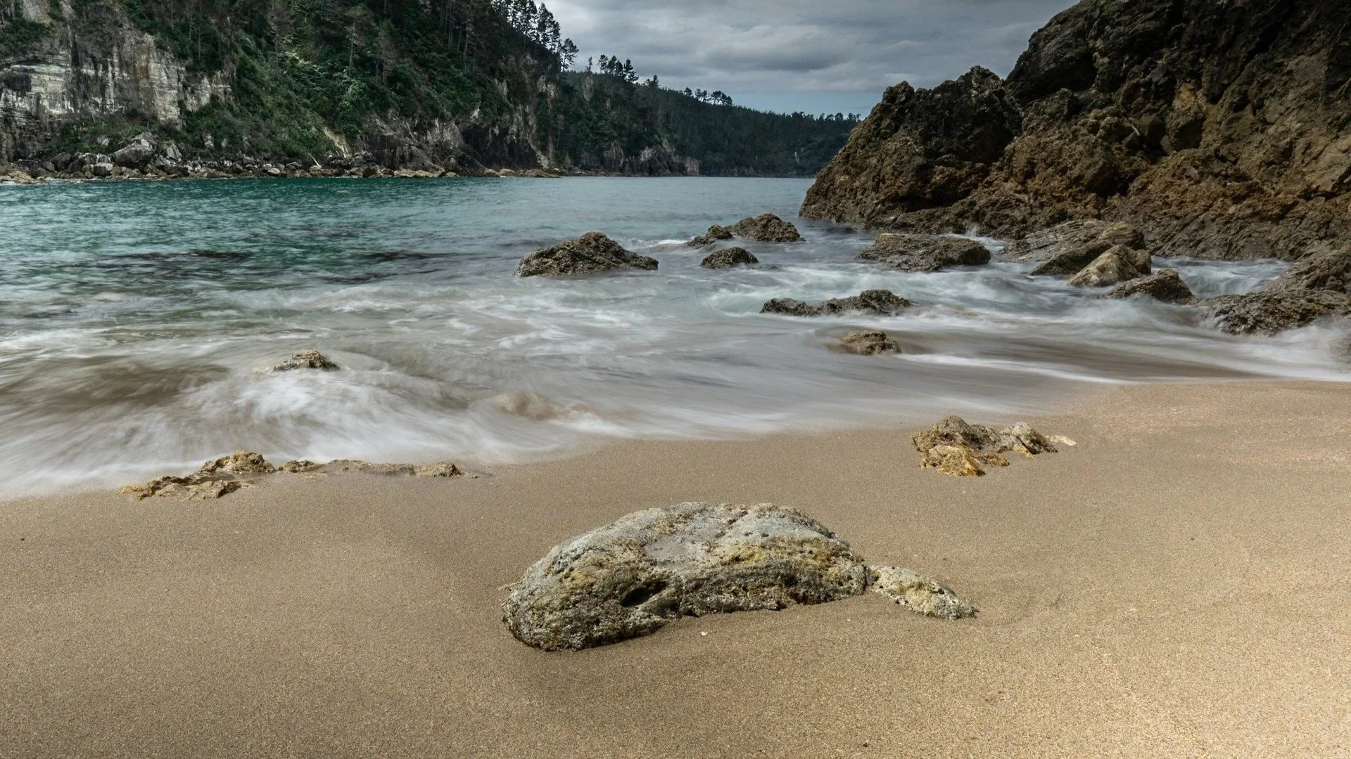 A rocky New Zealand coastline with soft waves washing over a sandy beach under an overcast sky.