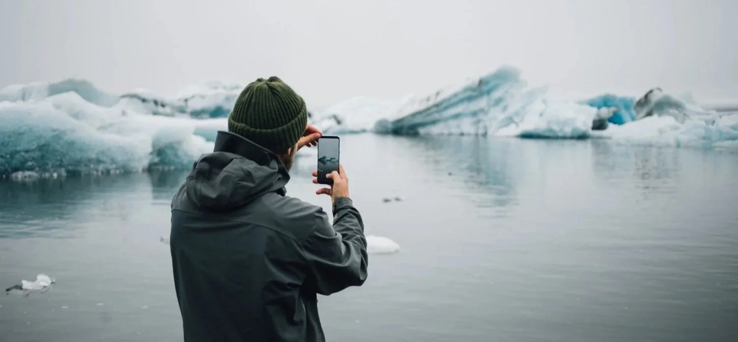 A person in a grey jacket and green beanie taking a smartphone photo of icebergs in a calm, misty glacial lagoon.