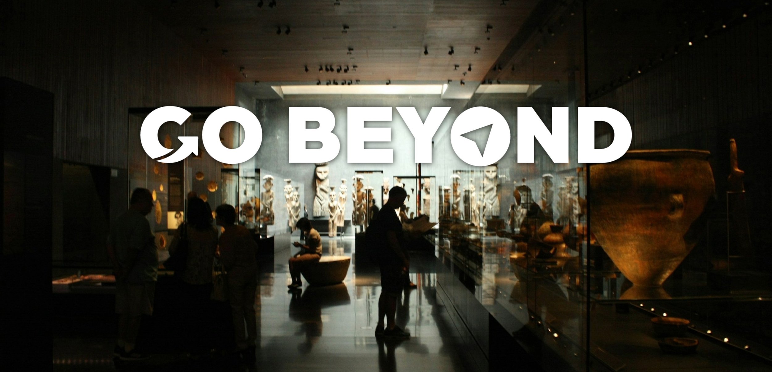 A photograph of people in a dimly lit museum gallery featuring large artifacts and statues, with the bold white text "GO BEYOND" overlaid in the center.