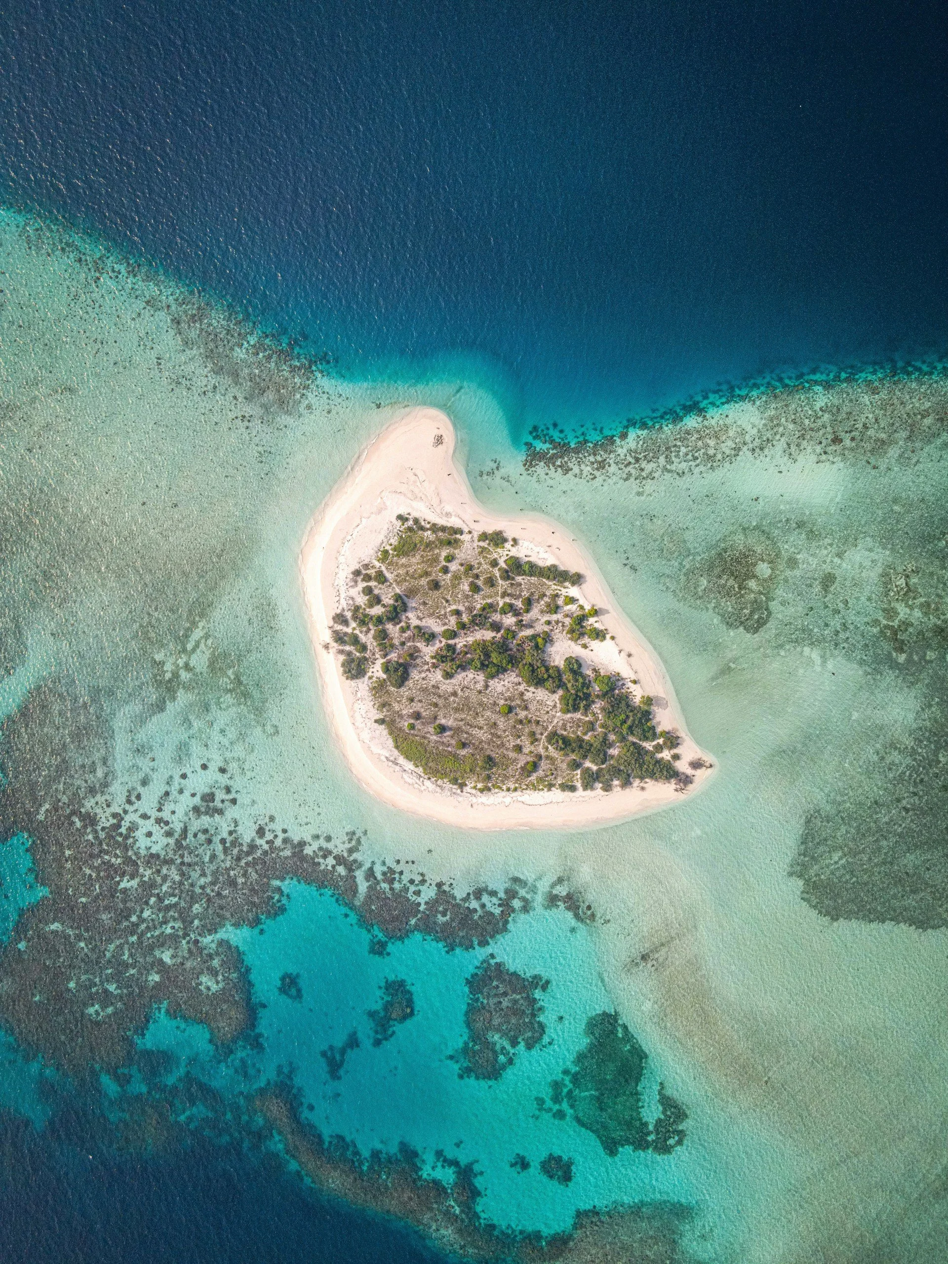 A high-angle aerial view of a small, lush tropical island surrounded by turquoise coral reefs and deep blue ocean water.