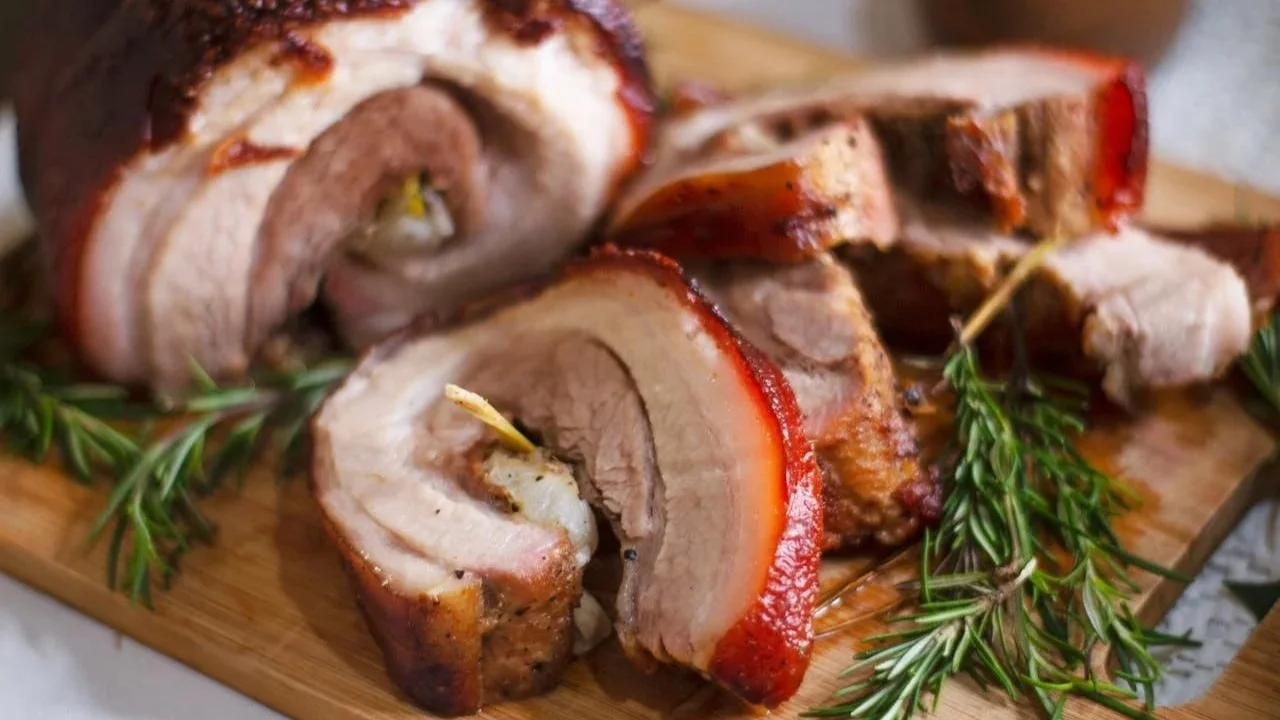 Slices of roasted pork belly lechon roll with crisp, reddish-brown skin arranged on a wooden cutting board next to fresh rosemary sprigs.